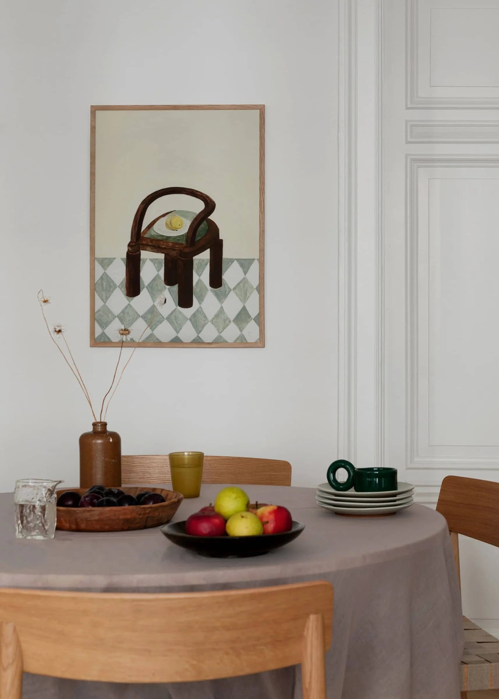 A round table with a beige tablecloth holds fruit, drinks, and green cups. Wooden chairs surround it; “Chair with Fruit” by Isabelle Vandeplassche (30x40) from The Poster Club decorates the white wall.