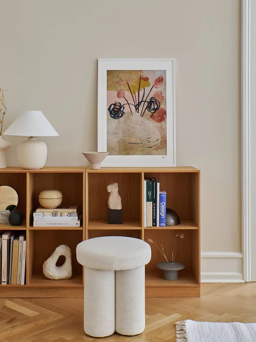 A minimalist living space with a wooden cubby shelf, books, ceramics, a white round stool, lamp, and The Poster Club Madelen Möllard - Flowers in Blue Vase (50x70) art print against a beige wall and herringbone wooden floor.