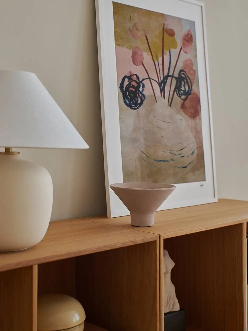 A minimalist wooden shelf displays a white lamp, a beige bowl, and The Poster Clubs Johannes Geppert - Blooming (50x70) print with abstract florals in muted tones, leaning against a light-colored wall.