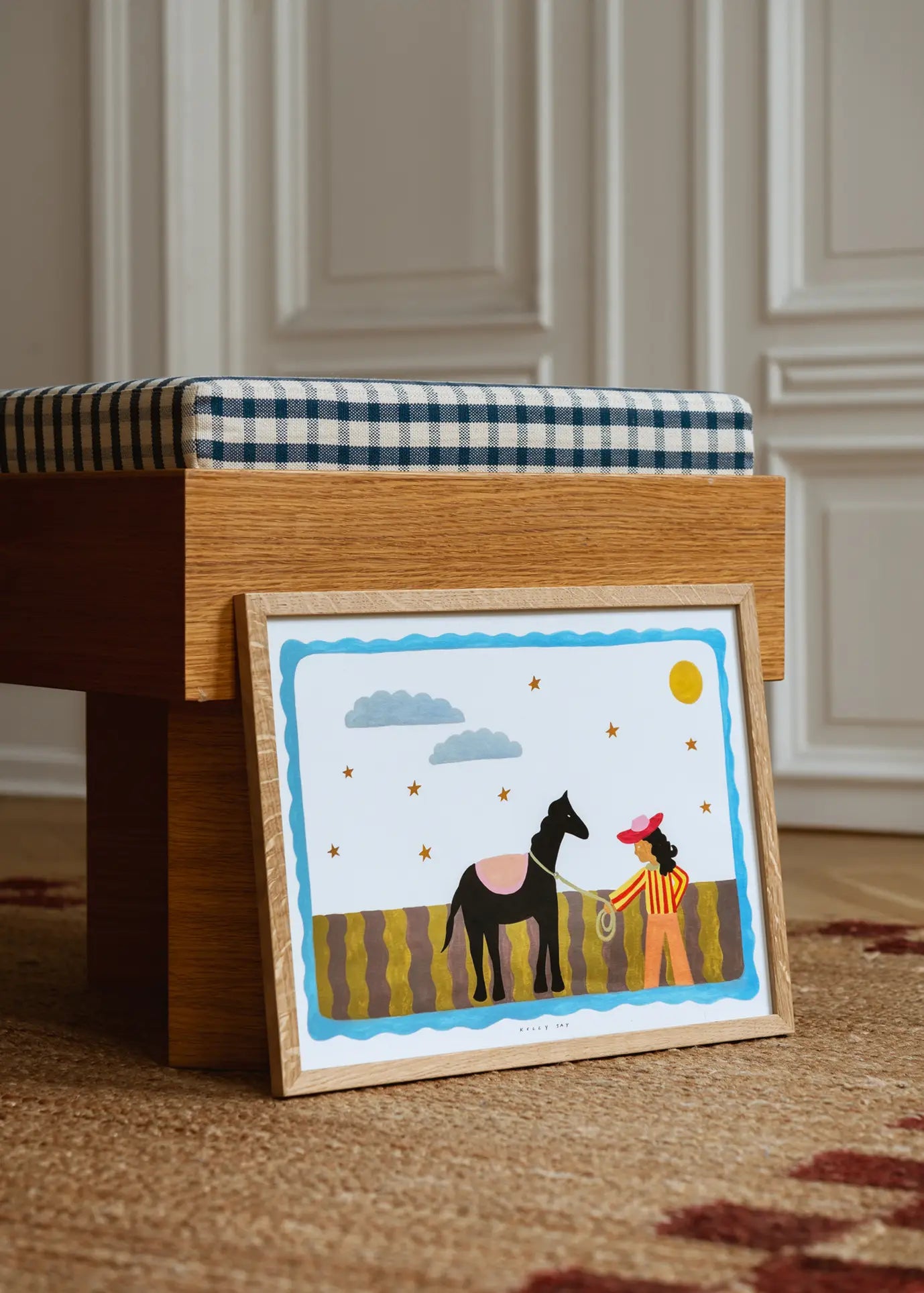 The Poster Club Kelly Jay - A Journey Begins (50x70) art print, inspired by Kelly Jay, features a person with a rope and black horse under a bright sky, framed and displayed against a bench in a stylish room with white paneled walls.