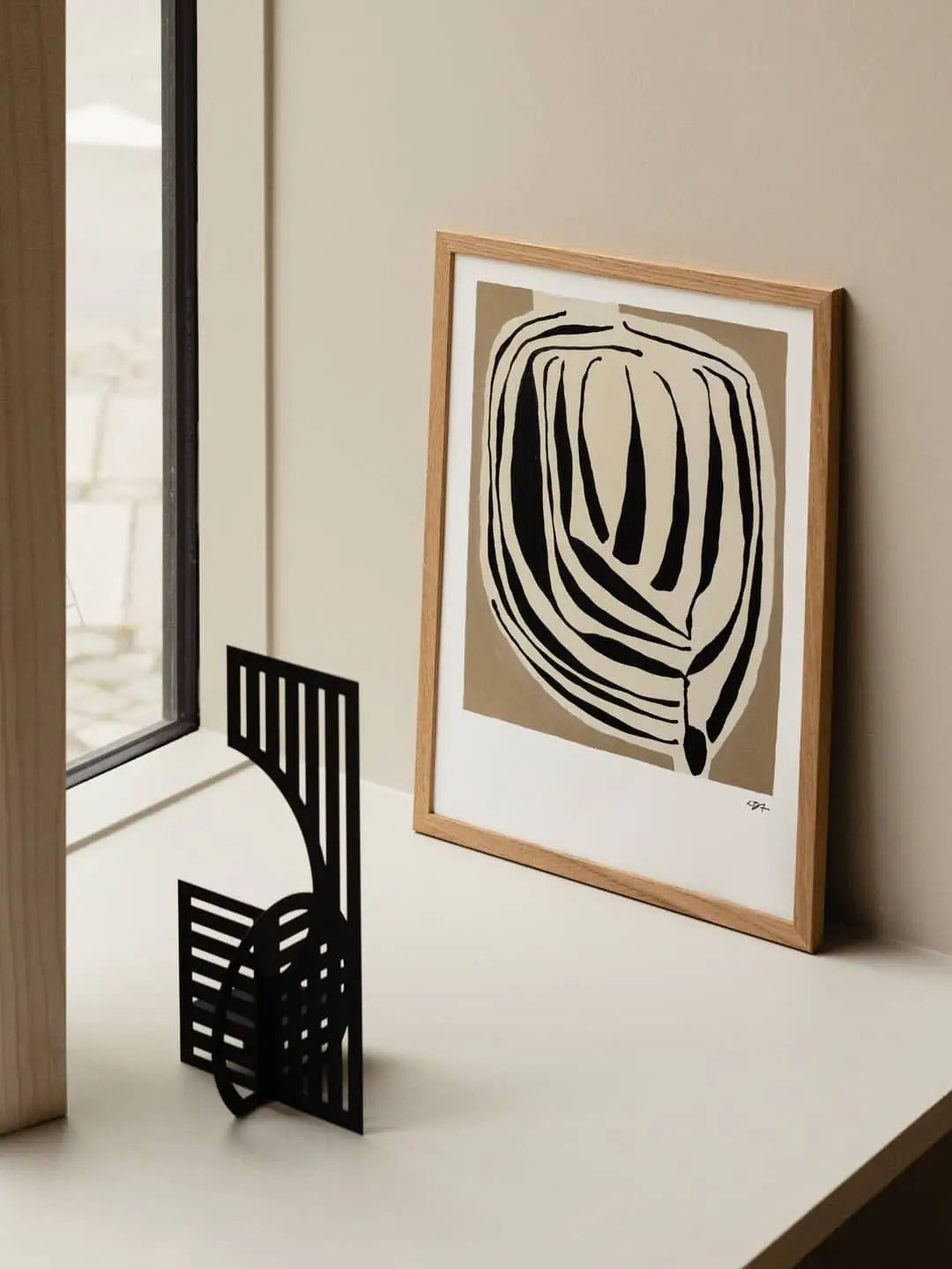 The Poster Club Berit Mogensen Lopez - Bloom (50x70) art print rests on a light desk against a beige wall, beside a black geometric metal sculpture near the window—an eye-catching piece from The Poster Club.