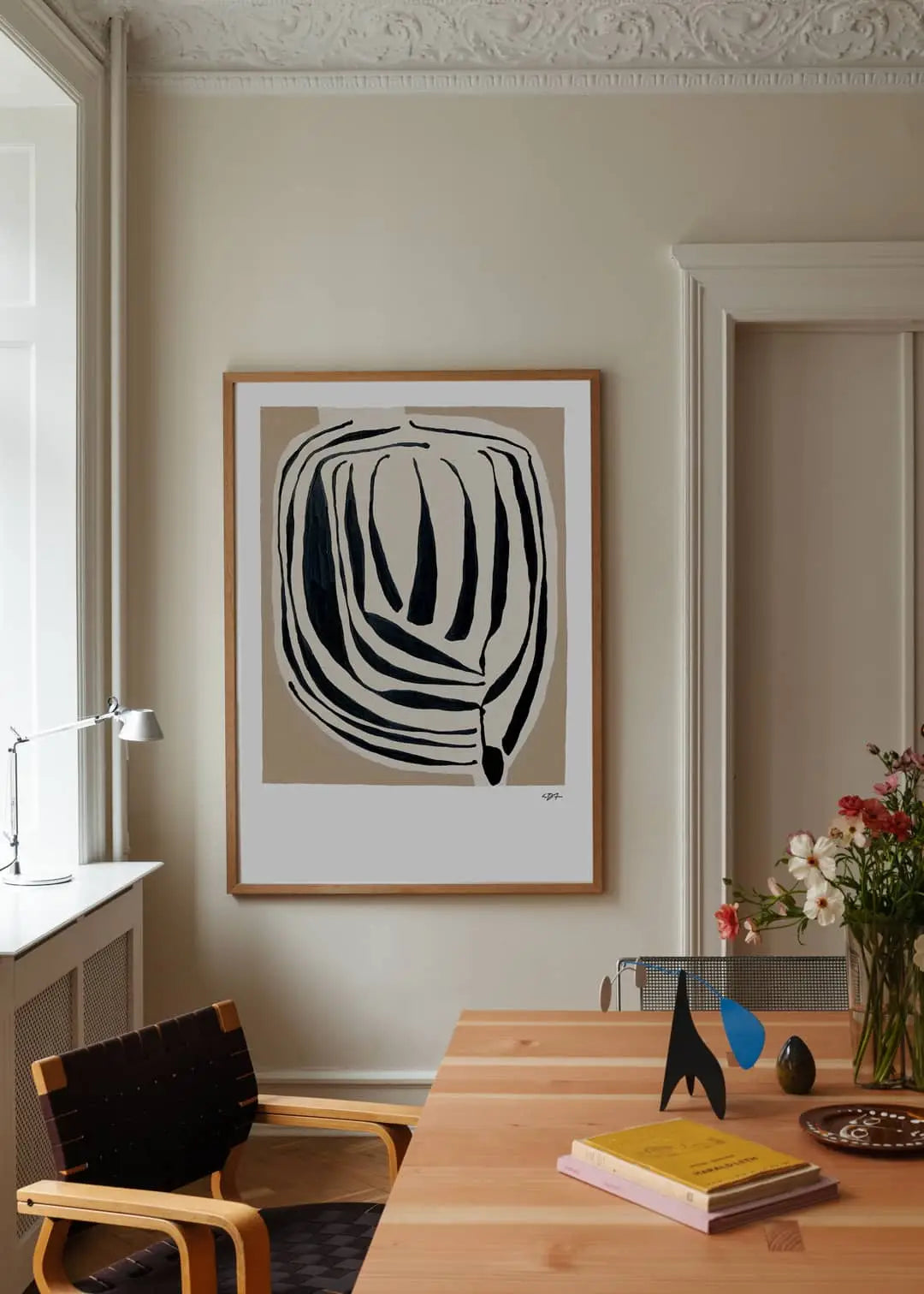 A minimalist home office features a wooden desk, modern chair, books, and flowers in a vase. Above hangs The Poster Club Berit Mogensen Lopez - Improvise (50x70) art print, with natural light streaming in from a nearby window.