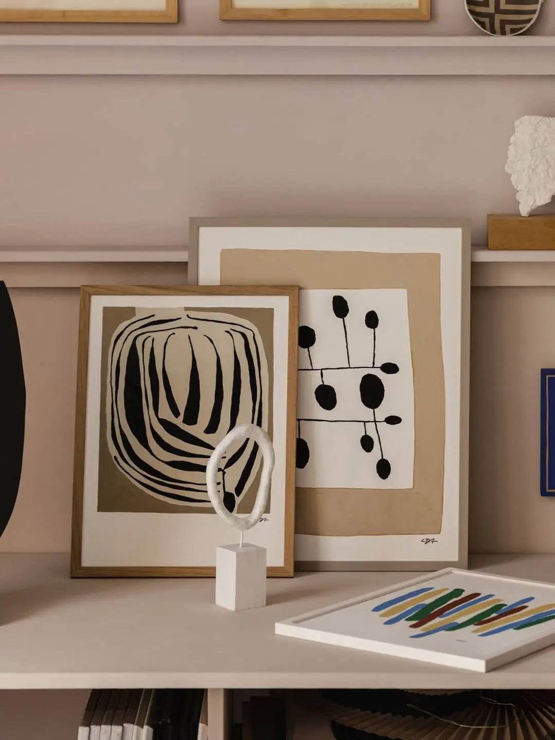 The Poster Clubs Bloom (50x70) by Berit Mogensen Lopez leans on a shelf with another black and white print; in front are a small white sculpture and a colorful book.