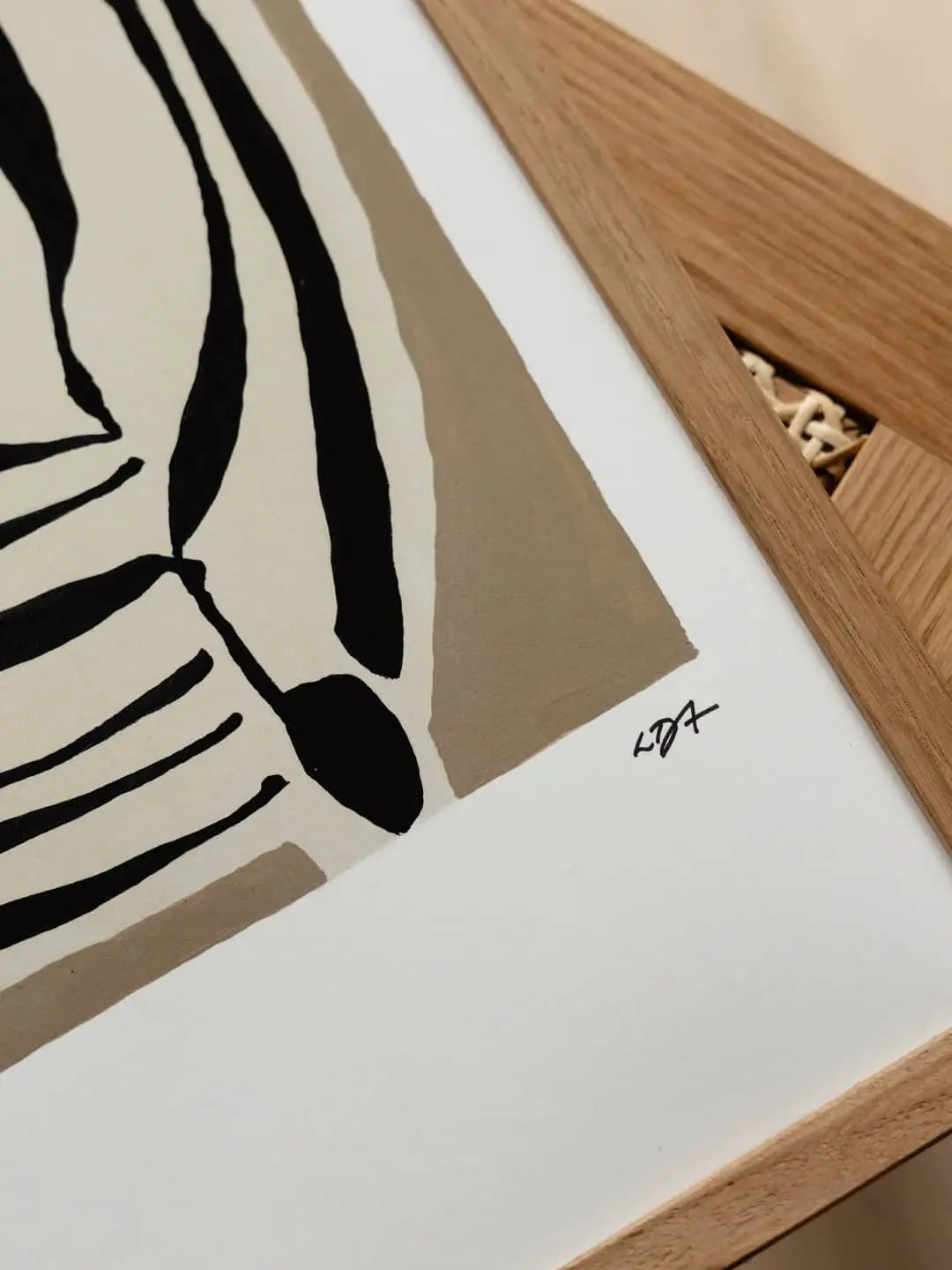 A close-up of The Poster Club Berit Mogensen Lopez - Improvise (50x70) art print, with abstract black and beige shapes, partially framed in light wood. The artist’s signature is in black ink on the white border. Brand: The Poster Club.