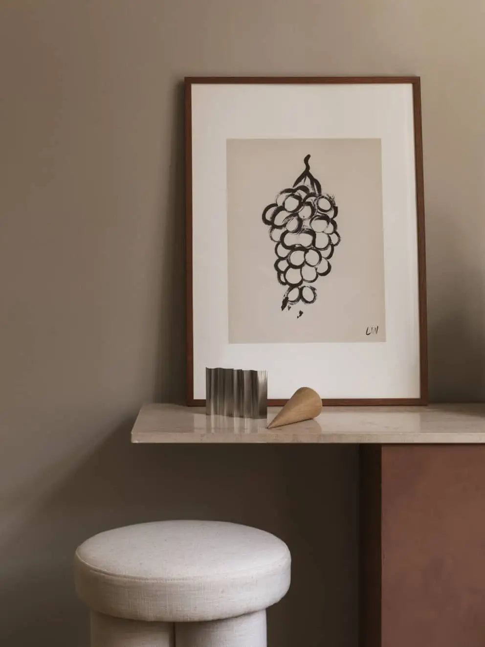 The Poster Club Lena Wigers - Grapes (50x70) giclée print features abstract grapes on a beige wall shelf with metallic and wooden accents. Below the shelf is a round, white upholstered stool from The Poster Club.