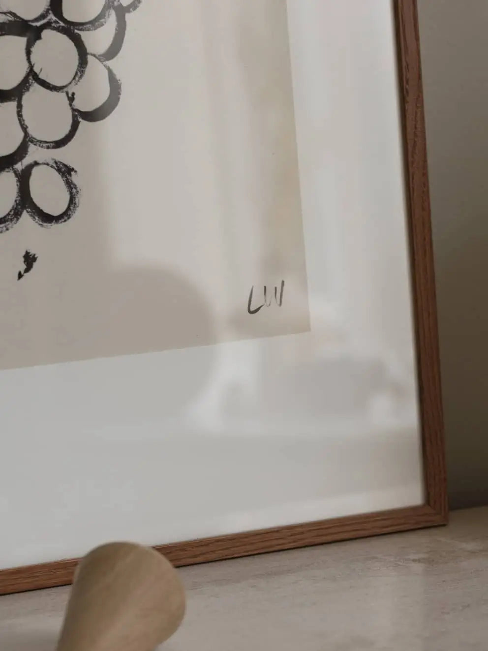 A close-up of The Poster Clubs Madelen Möllard - Flowers in Blue Vase (50x70) art print in a wooden frame, featuring a handwritten “Lill” signature in the corner. A blurred foreground object is visible.