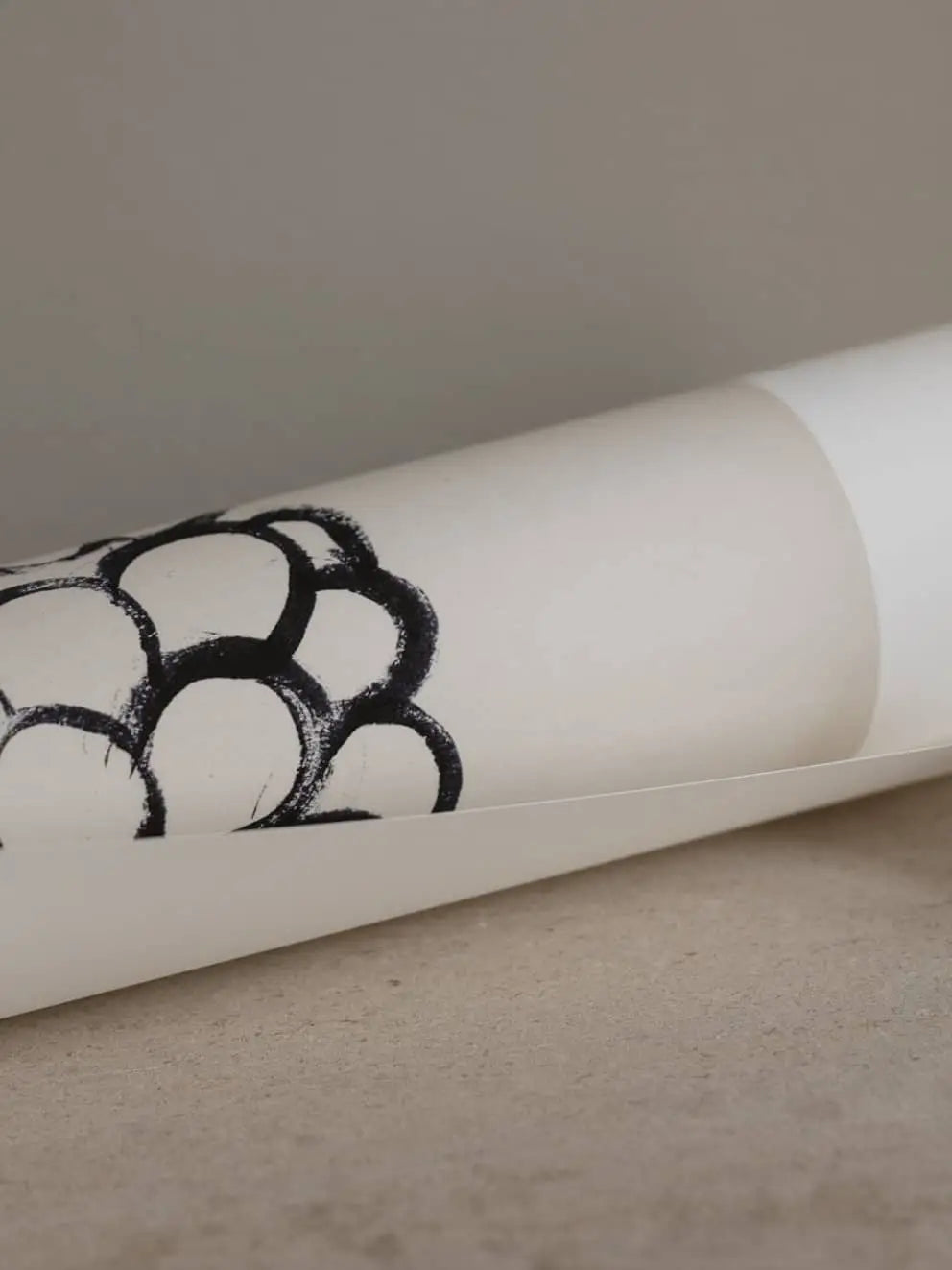 A rolled-up sheet of white paper with a black abstract circular pattern, from The Poster Club Madelen Möllard - Flowers in Blue Vase (50x70) art print by The Poster Club, rests on a neutral-colored surface.