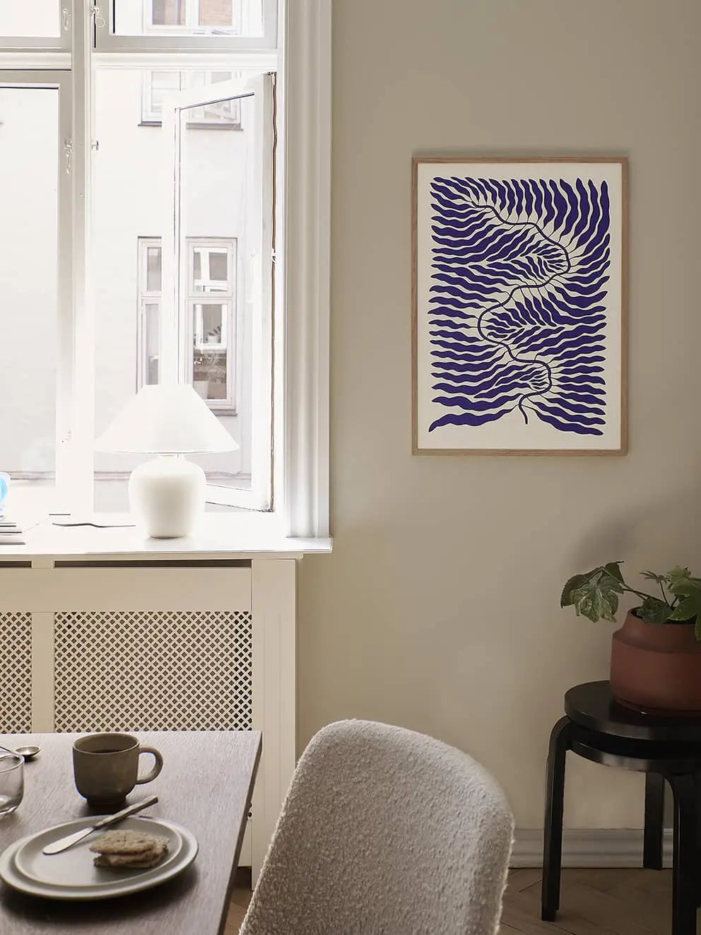 A cozy dining area features a light wooden table, gray chair, and natural light. The Poster Clubs Linnéa Andersson - Little Blue (50x70) abstract blue leaf art hangs on the wall; a white vase lamp sits on the windowsill.