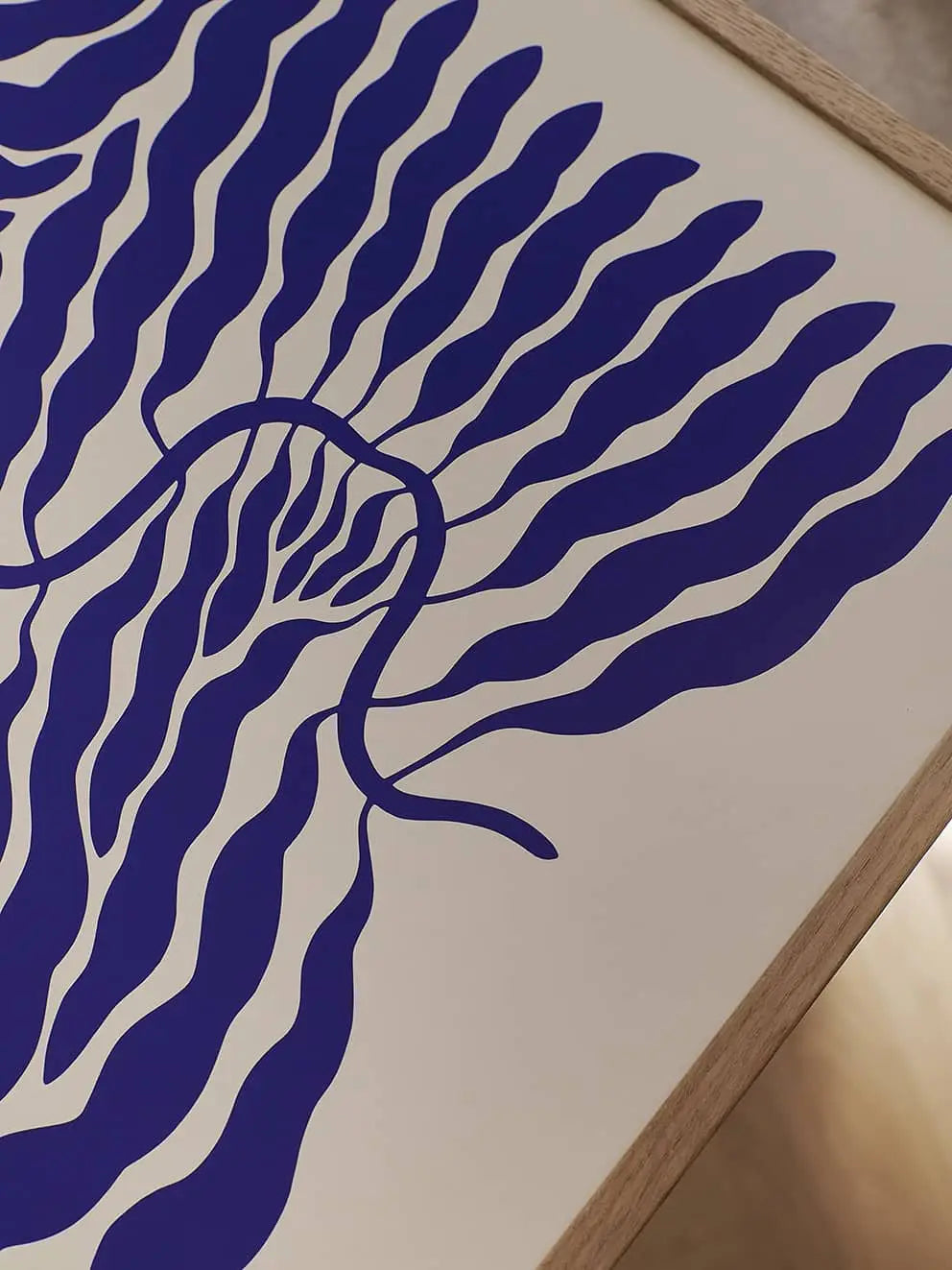 The Poster Club Linnéa Andersson - Little Blue (50x70) art print features wavy blue lines radiating from a central shape on a cream background, evoking stylized leaves or water. Framed in light wood, its inspired by Andersson’s signature leaf motif.