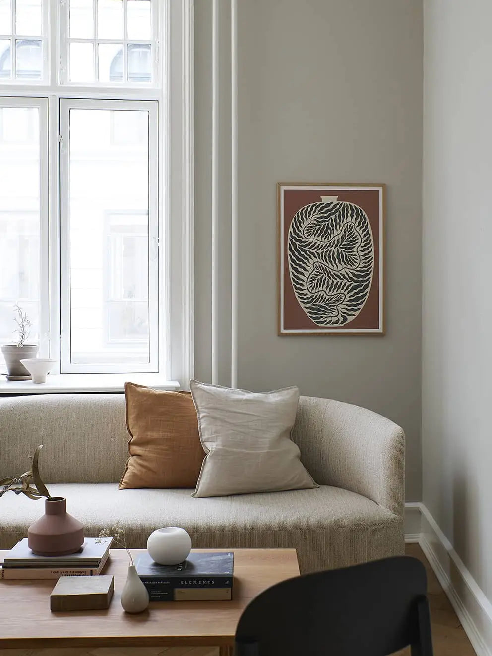 A minimalist living room with a curved beige sofa, tan and white pillows, a wooden coffee table with decor, Madelen Möllards Flowers in Blue Vase (50x70) art from The Poster Club on the wall, and a large window letting in natural light.