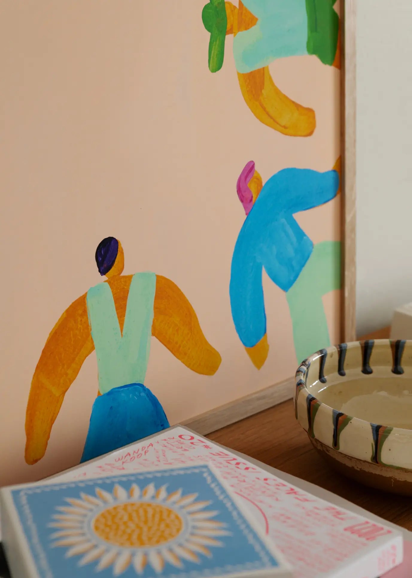 The Poster Club Lise Uhrskov - On The Edge (30x40) giclée print shows colorful abstract human figures and is displayed on a shelf with a ceramic bowl and a book featuring a sun illustration.