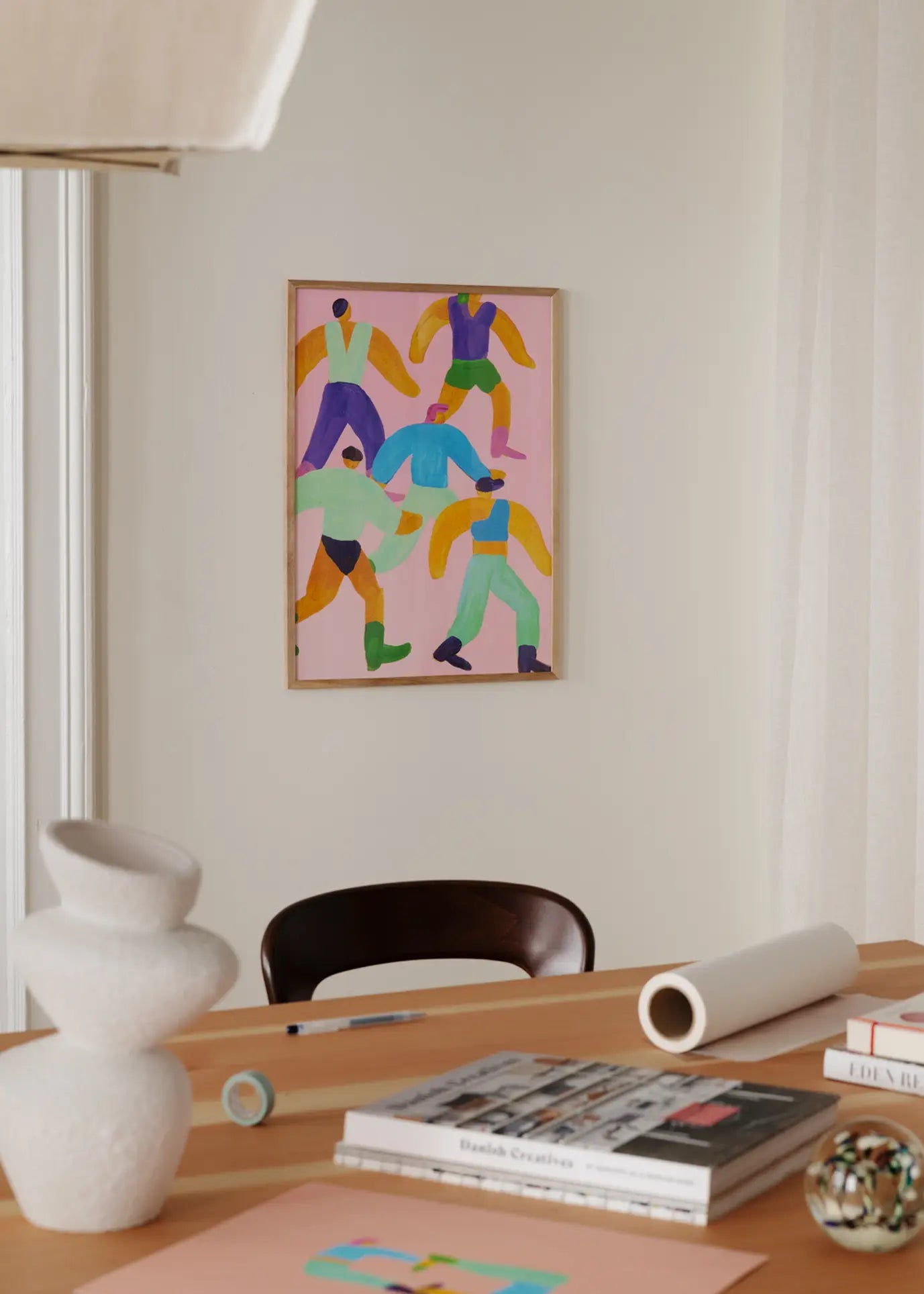 A modern workspace with a wooden desk topped with books, papers, a sculptural vase, and a decorative sphere, featuring The Poster Club Lise Uhrskov - Walk The Walk (50x70) fine art print hanging on the beige wall.