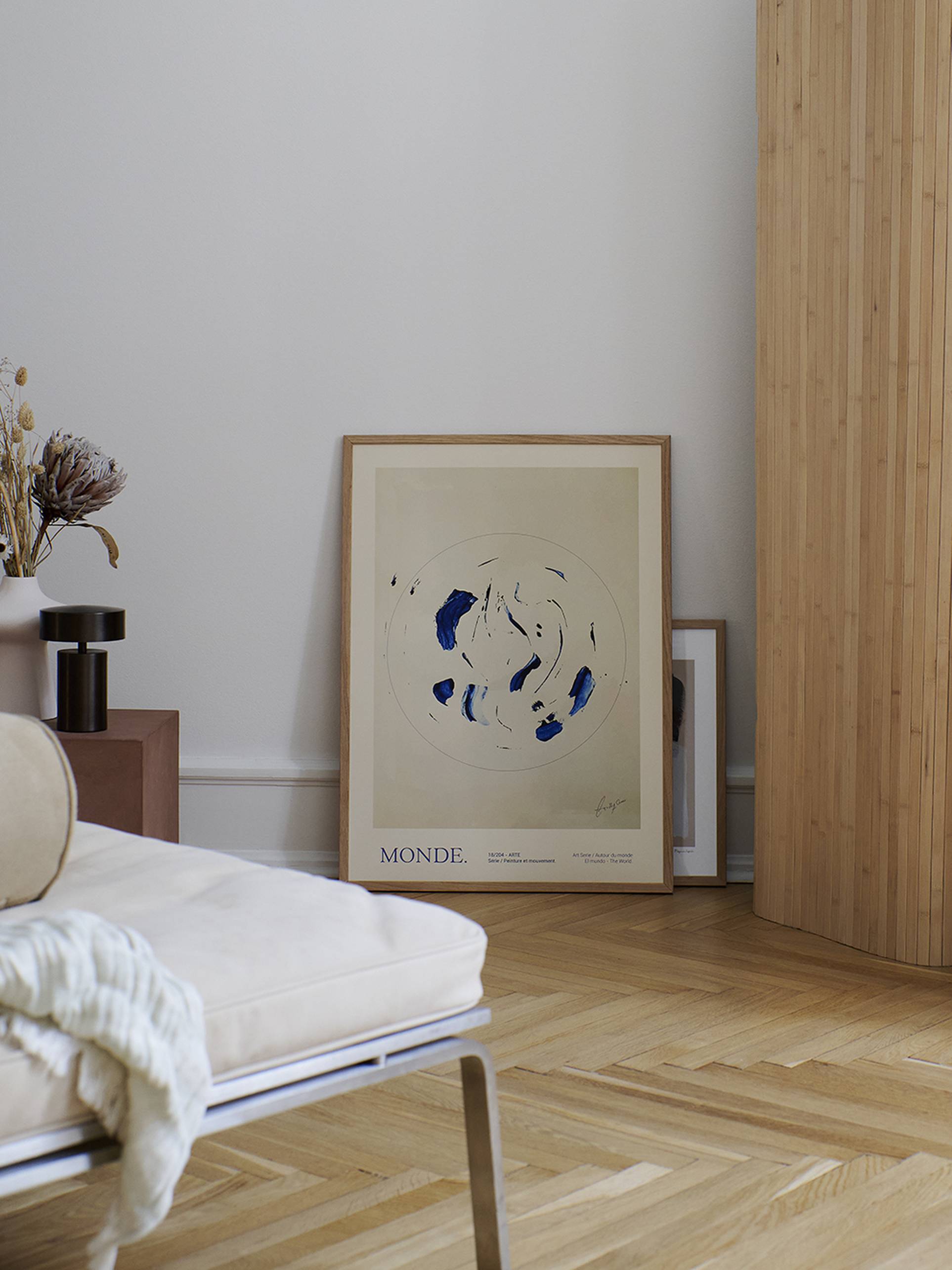 A minimalist room corner displays The Poster Club Lucrecia Rey Caro - Le Monde (50x70) art print leaning against the wall on a wooden floor, accompanied by a round beige chair, folded white blanket, dried flowers, and a small brown table lamp.