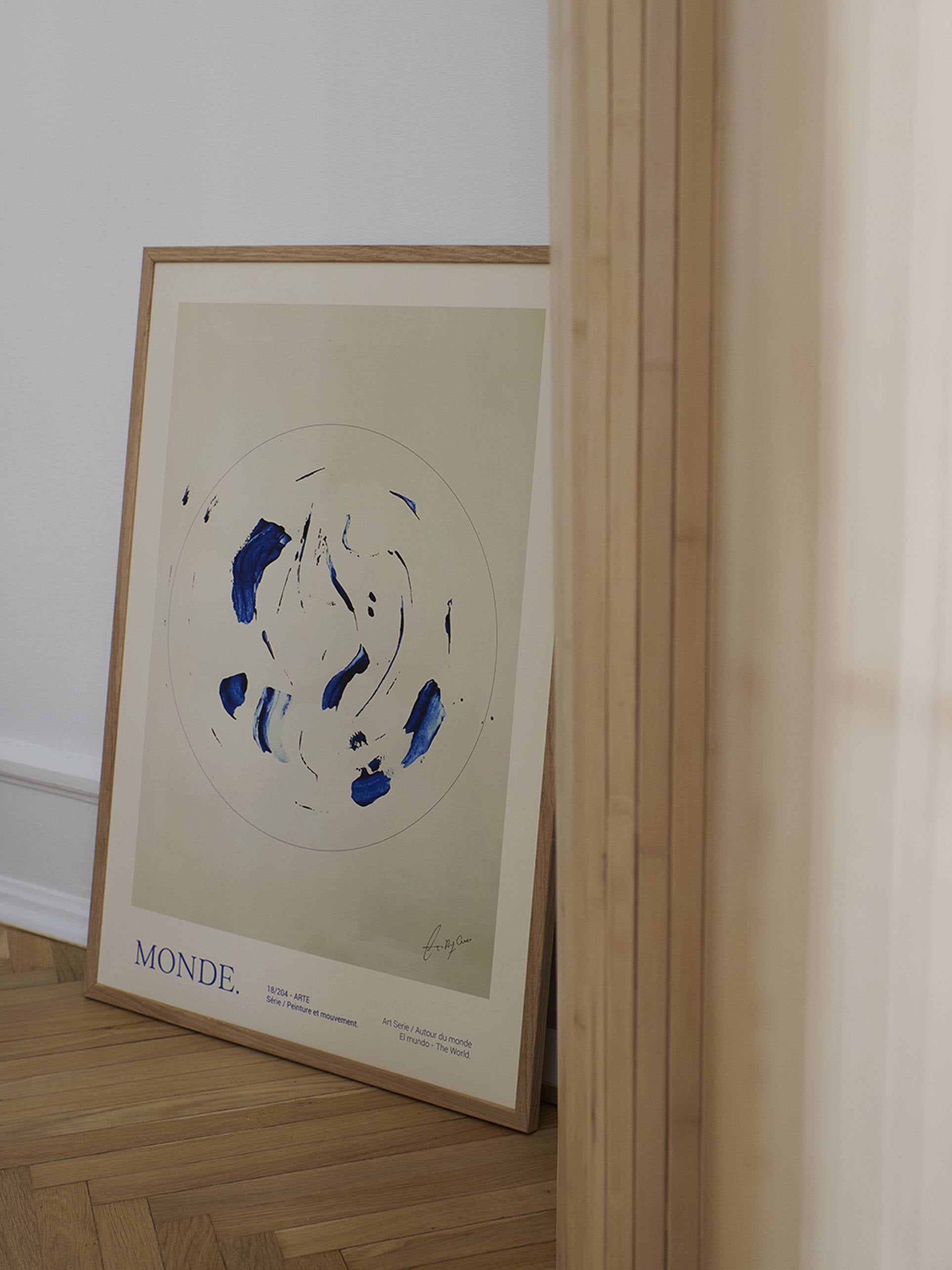 The Poster Club Lucrecia Rey Caro - Le Monde (50x70) art print features blue and black brushstrokes on a beige background, leaning against a white wall on a wooden floor, partially hidden by a light vertical panel. Available at The Poster Club.