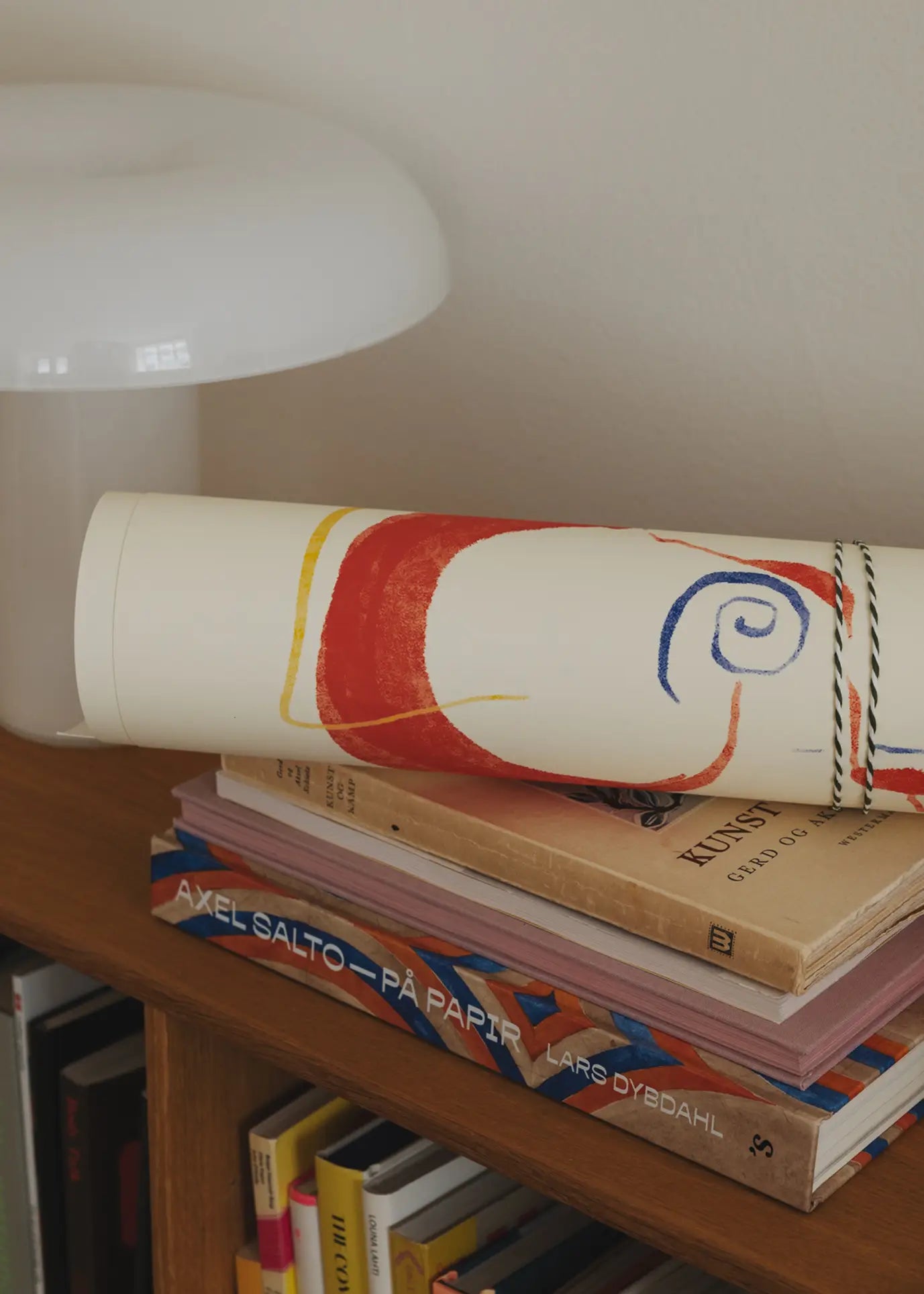 A rolled-up Lucrecia Rey Caro – Nos Encontramos (30x40) art print from The Poster Club sits on stacked books, including AXEL SALTO – PÅ PAPIR, atop a wooden shelf next to a white mushroom-shaped lamp.