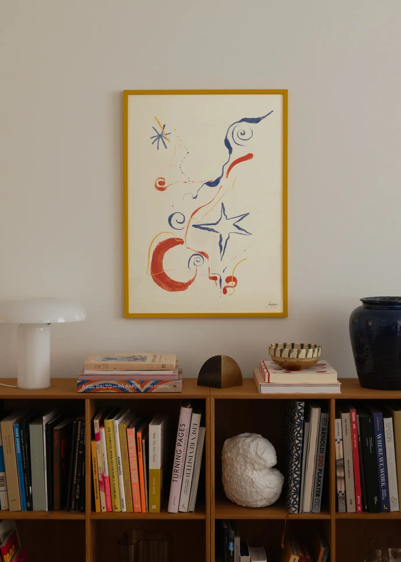 A bookshelf holds assorted books and decor beneath The Poster Clubs Lucrecia Rey Caro - Nos Encontramos (30x40) art print. On the shelf are a white lamp, bowl, and blue vase. The print features bold red, blue, and orange shapes.