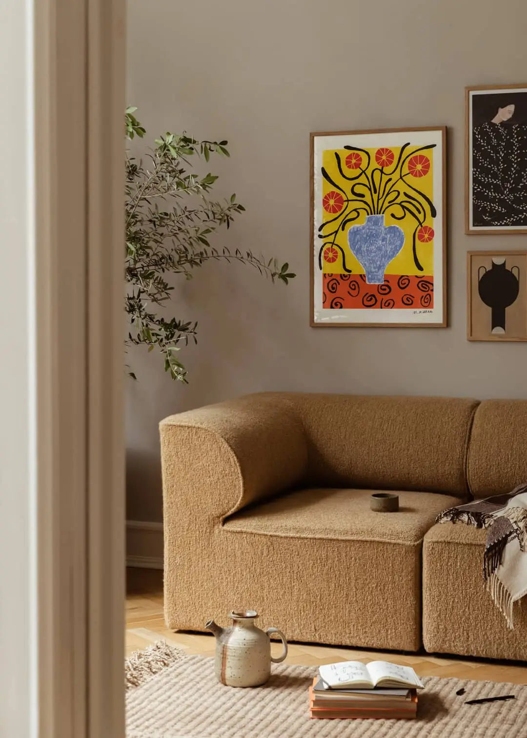 A cozy living room features a tan textured sofa, leafy plant, rug, open book, teapot and cup on the floor. The Poster Club’s Madelen Möllard - Flowers in Blue Vase (50x70) print adds color to the light-toned wall.