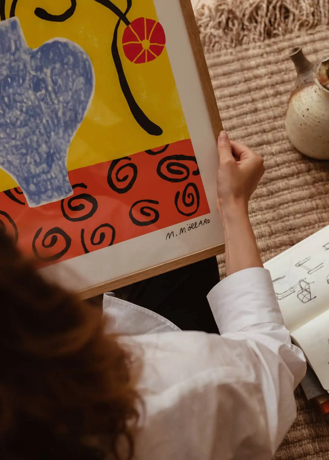 A person holds The Poster Club Madelen Möllard - Flowers in Blue Vase (50x70) art print while sitting on a textured rug near a ceramic vase and open sketchbook—a scene inspired by The Poster Club.