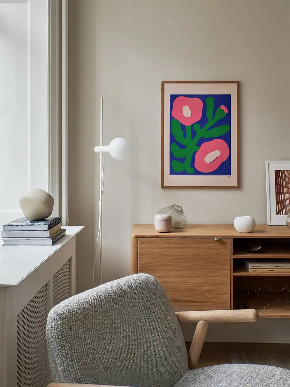 A modern living room styled with a light gray armchair, wood sideboard, books, ceramic decor, and Flowers in Blue Vase by Madelen Möllard (50x70) from The Poster Club. Neutral tones and minimalist design enhance the space.