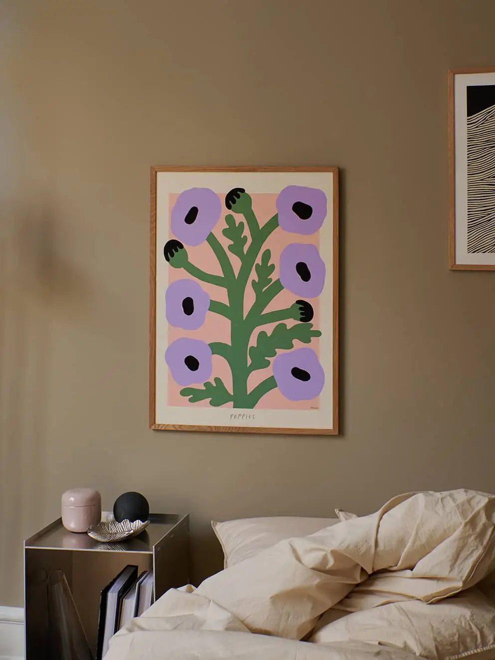The Poster Club Madelen Möllard - Purple Poppies (50x70) print hangs on a beige wall above an unmade bed, with a small metal side table nearby displaying decorative items.
