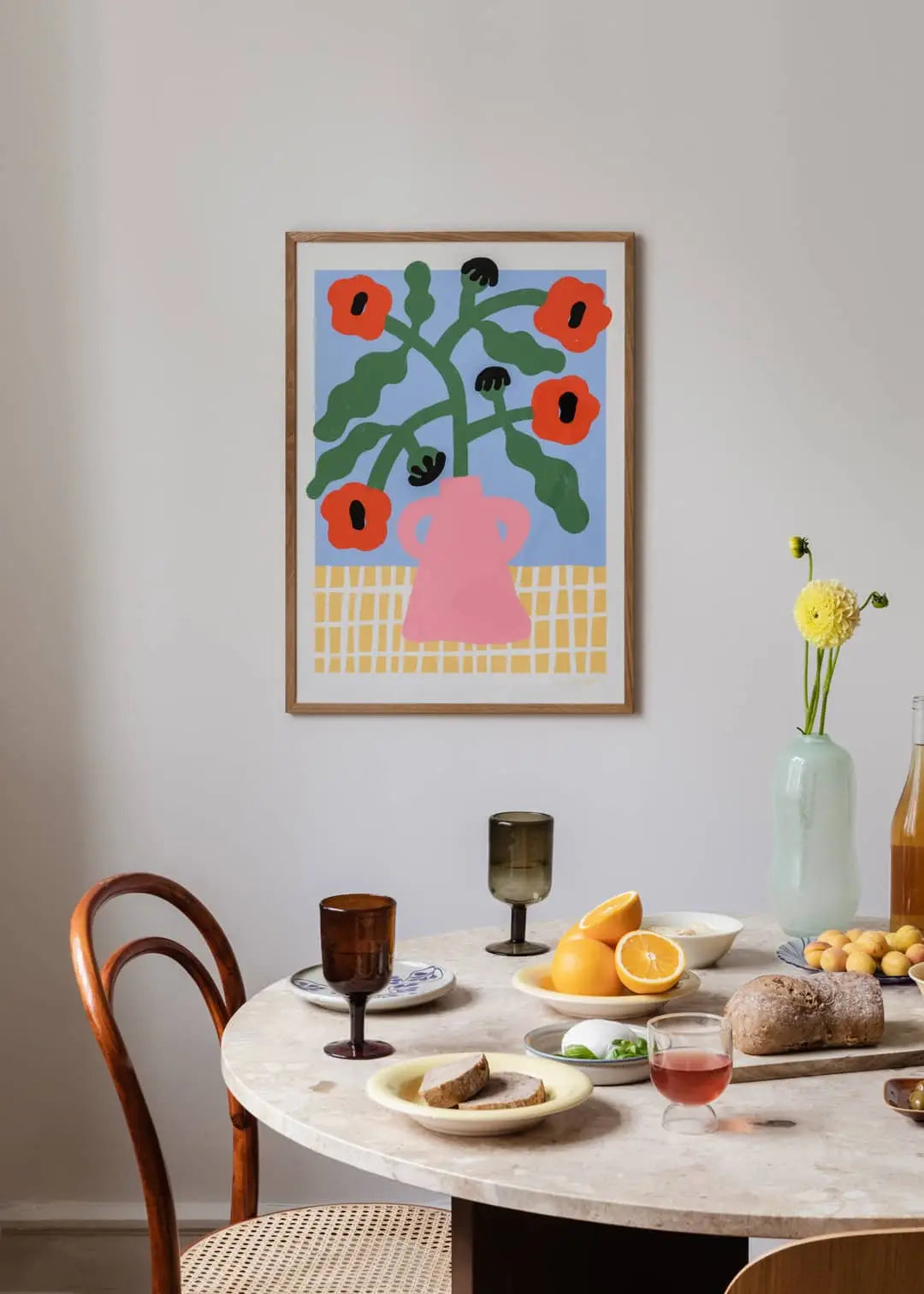 A round dining table set with bread, fruit, and drinks sits in a cozy room. Above it hangs The Poster Club Madelen Möllard - Red Poppies in Pink Vase (50x70) print, showing red poppies and green leaves in a pink vase on a blue background.