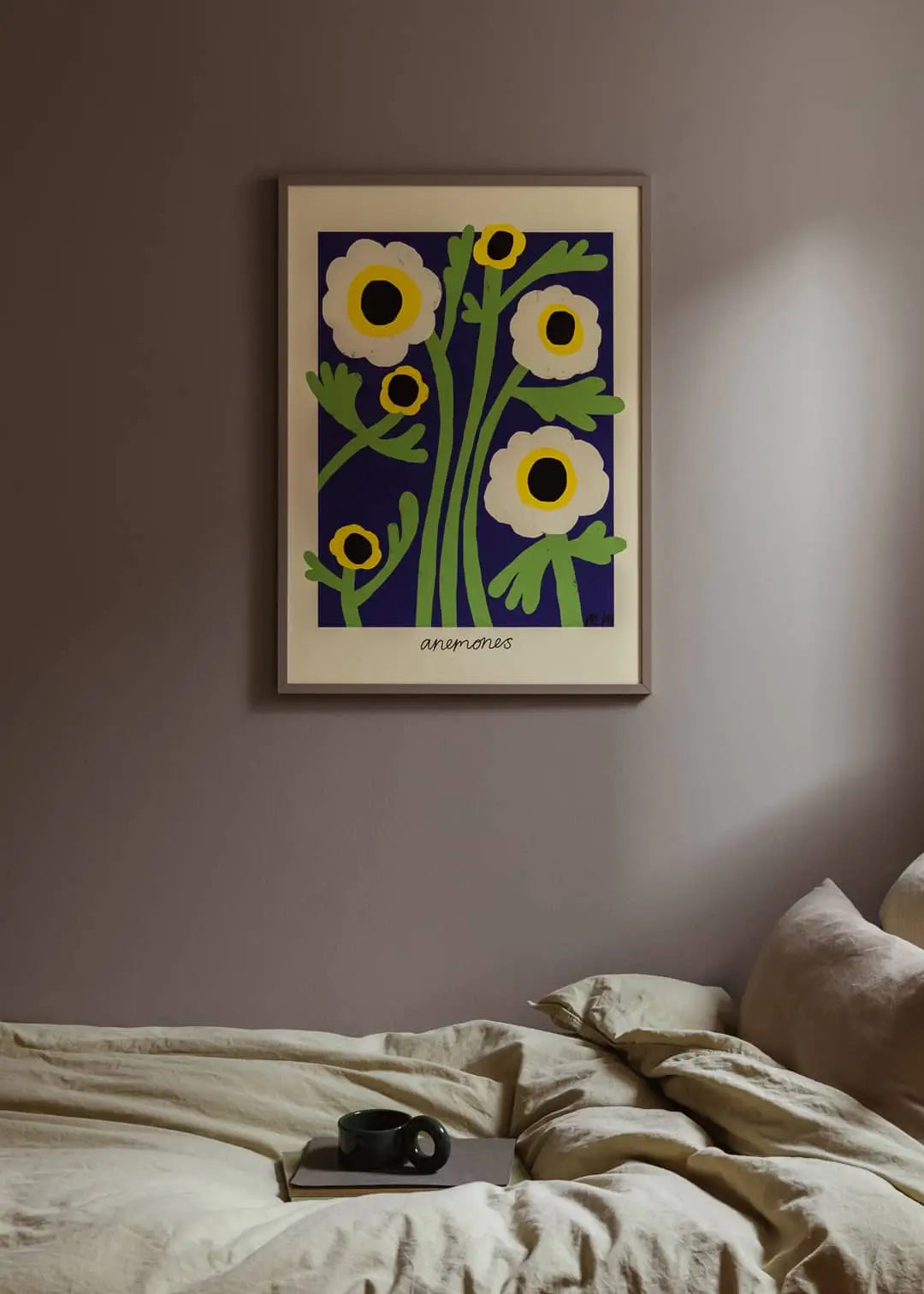 The Poster Clubs Madelen Möllard - Yellow Anemones (50x70) art print in green, white, and yellow hangs above a beige bed, with a dark mug on the bedding and soft, warm lighting illuminating the taupe wall.