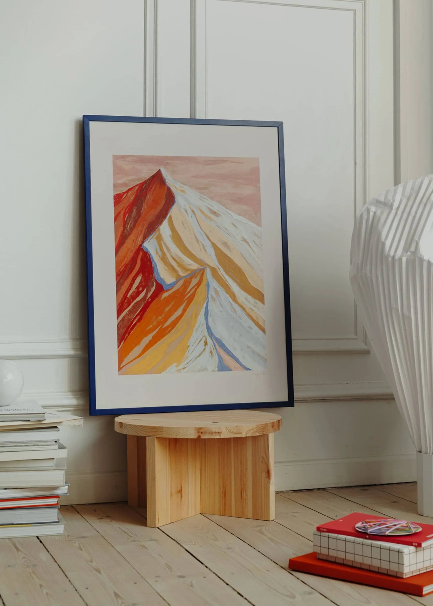 The Poster Clubs Mandy Maria - Cuillin Ridge VII (30x40) framed giclée print, with abstract red, orange, and white peaks, leans against a white wall on a small wooden stool beside stacked books and a pleated lamp.