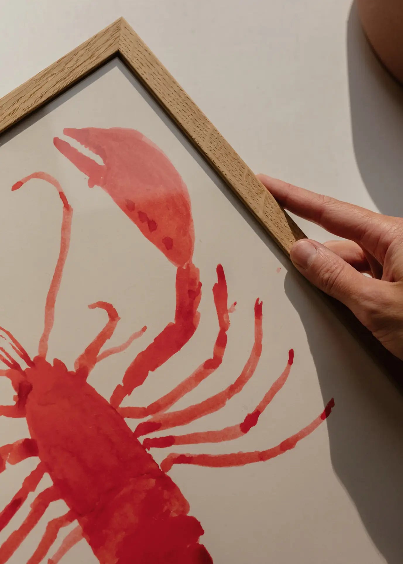 A hand holds the wooden frame of The Poster Club Marina García Fernández - Langosta (30x40) art print, featuring a vibrant red lobster on white paper. Available from The Poster Club.