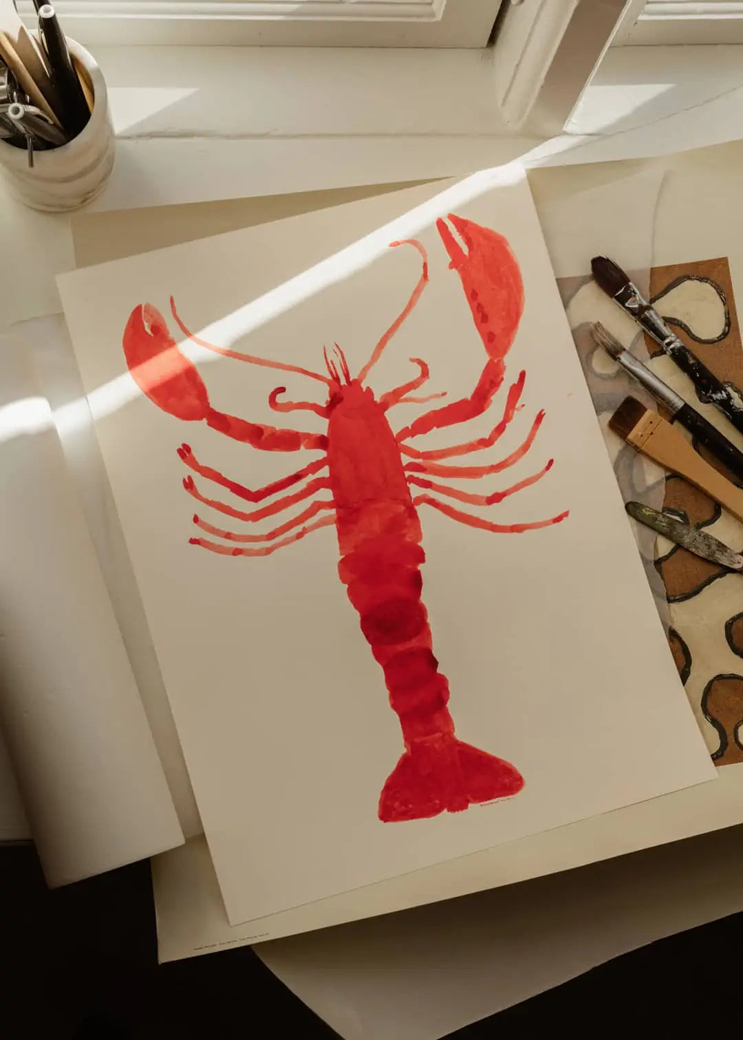 A floral art print, The Poster Club Berit Mogensen Lopez - Bloom (50x70), by The Poster Club, is displayed on a table with art supplies and brushes, illuminated by sunlight streaming through a nearby window.