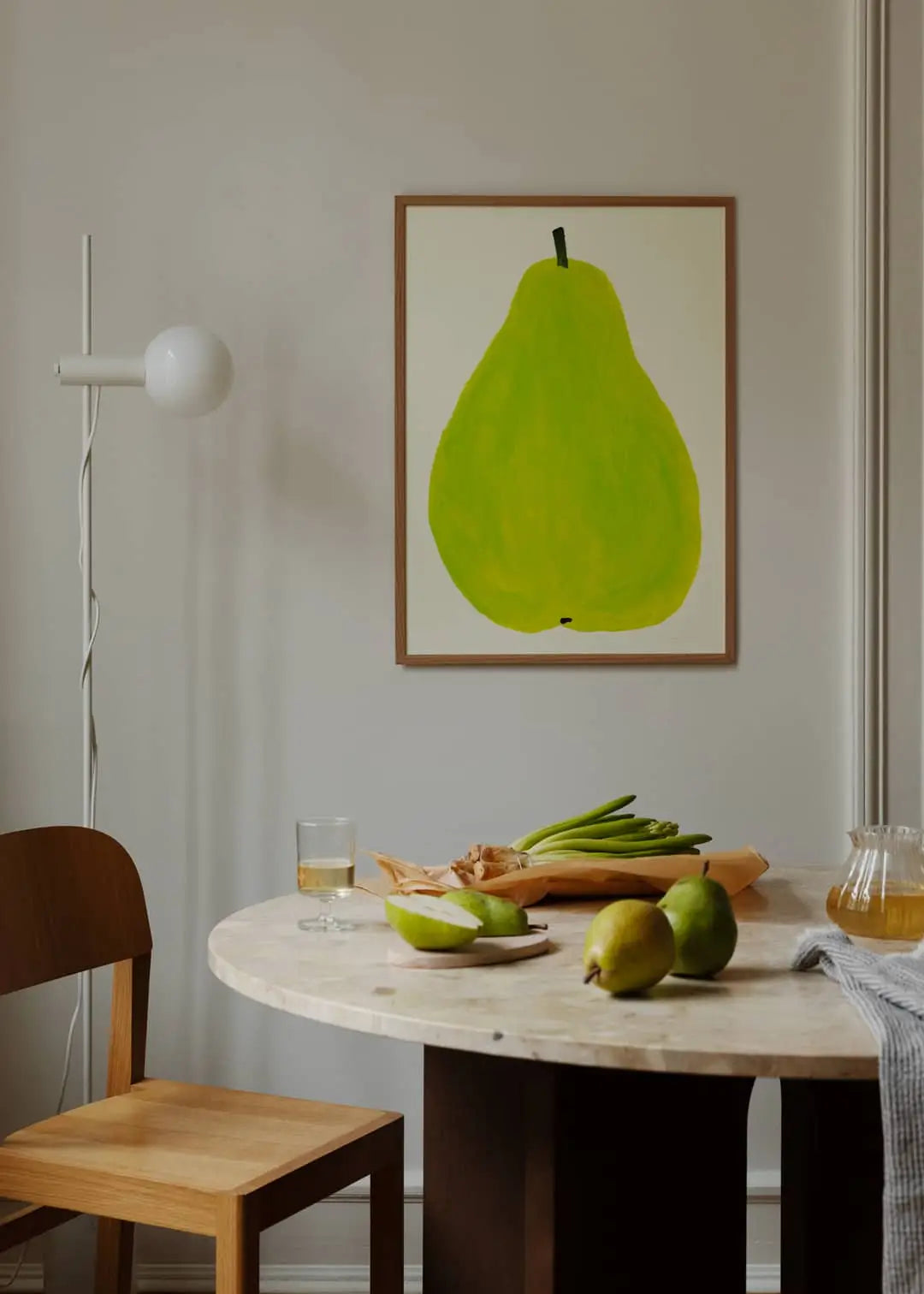 A minimalist dining area has a round table with pears, a glass of juice, and green onions. On the light wall hangs The Poster Club Marina García Fernández - Pera Limonera 1 (50x70) art print featuring a large green pear.