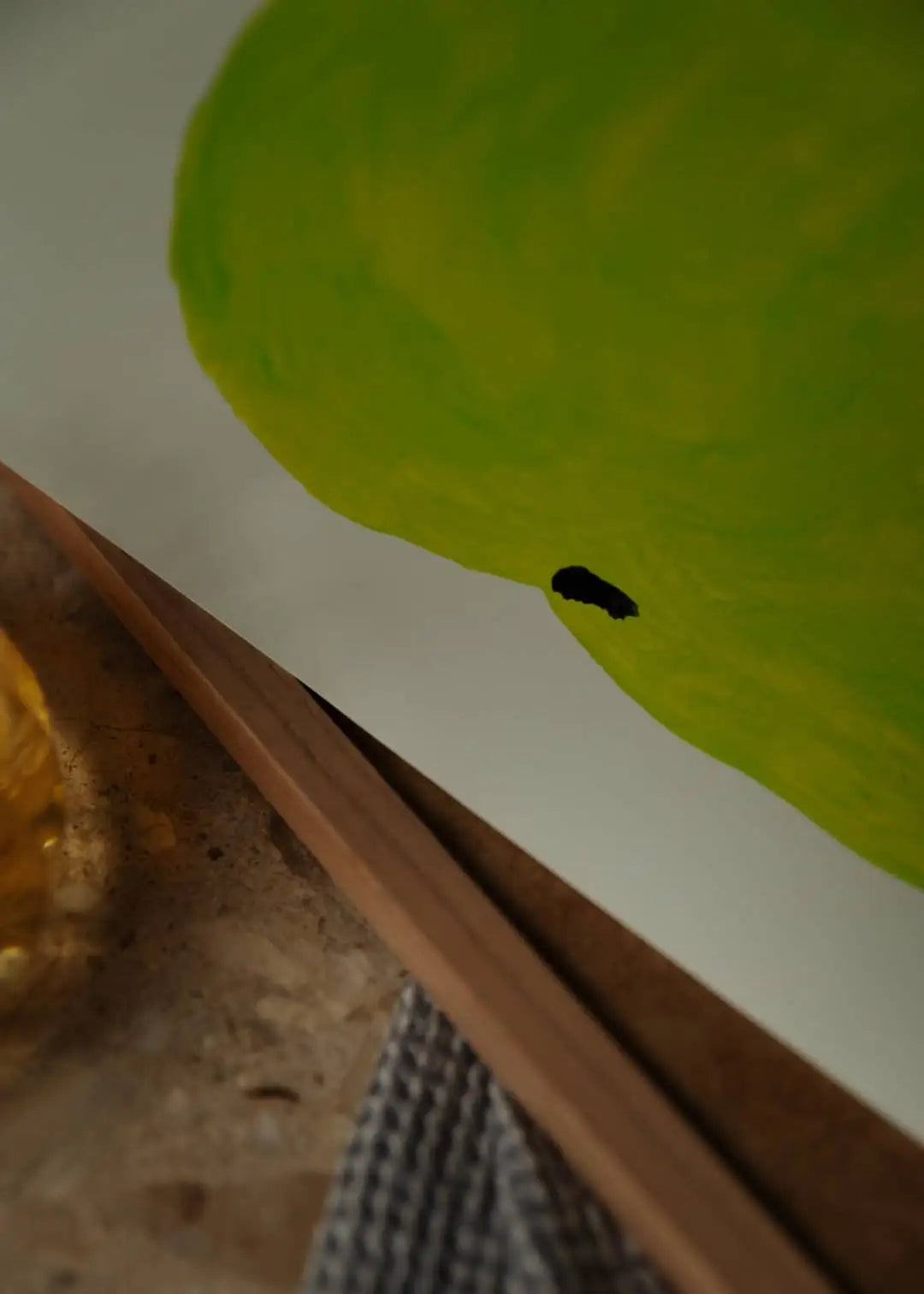 A close-up of The Poster Club Berit Mogensen Lopez - Bloom (50x70) art print shows part of a green pear with a small black spot, next to a wooden frame, textured gray cloth, and glass object in an elegant The Poster Club style.