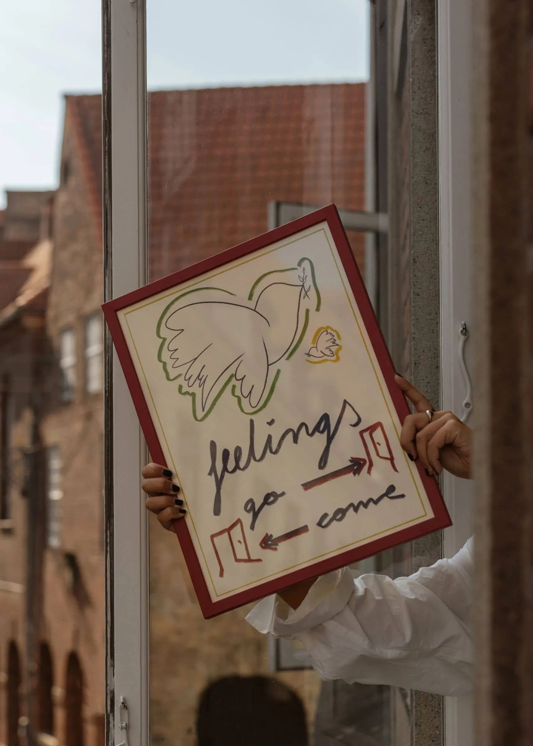 A person holds The Poster Clubs Marta Leyva - Feelings (30x40) giclée print, featuring a dove with an olive branch above the words feelings go come and two red arrows; brick buildings form a Mediterranean-inspired backdrop.