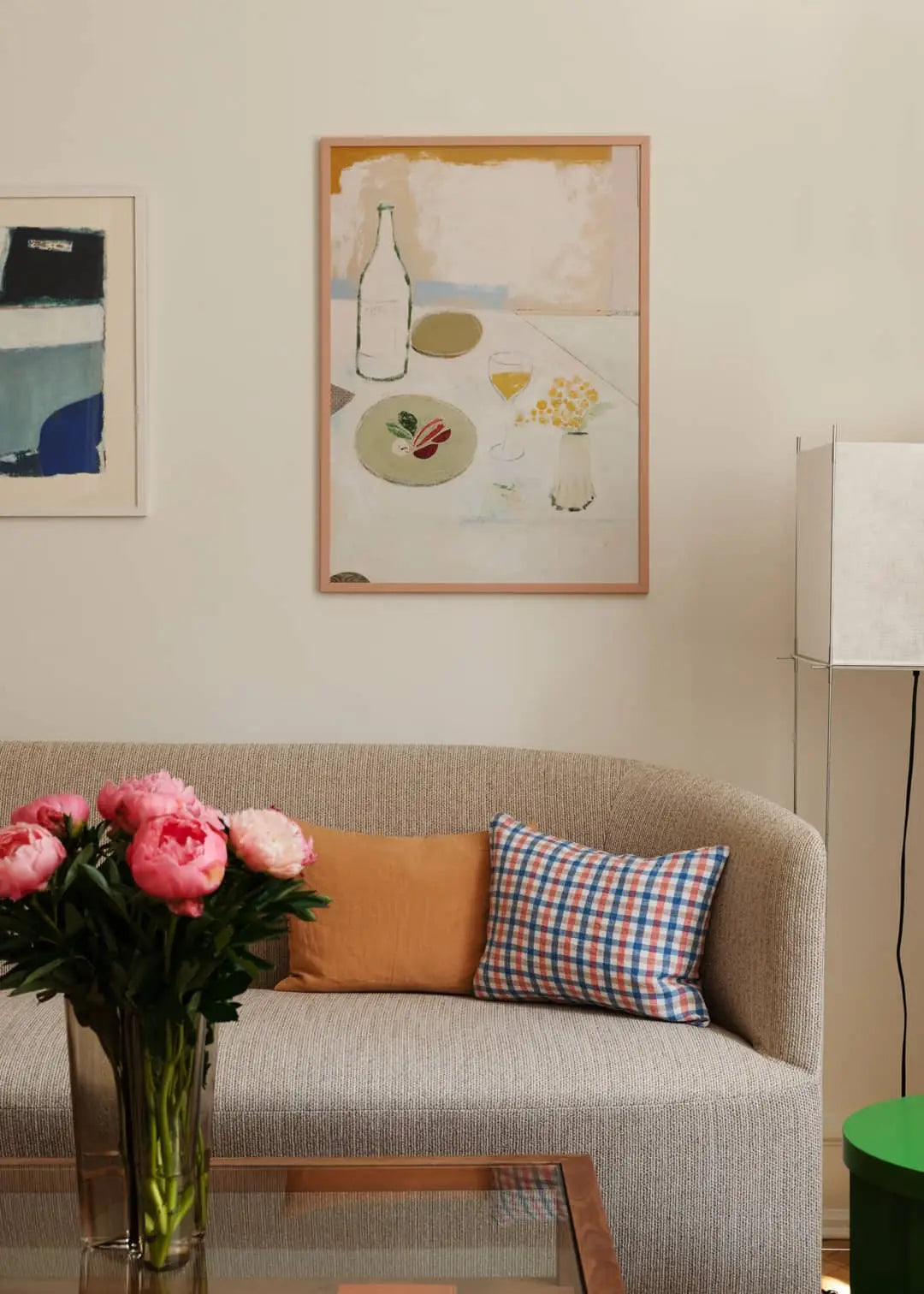 A cozy living room with a textured beige sofa, vintage textiles, mustard and plaid pillows, a glass vase of pink peonies on a glass coffee table, and The Poster Club Max Ososki - Fleurs Jaunes (50x70) art print on the wall.