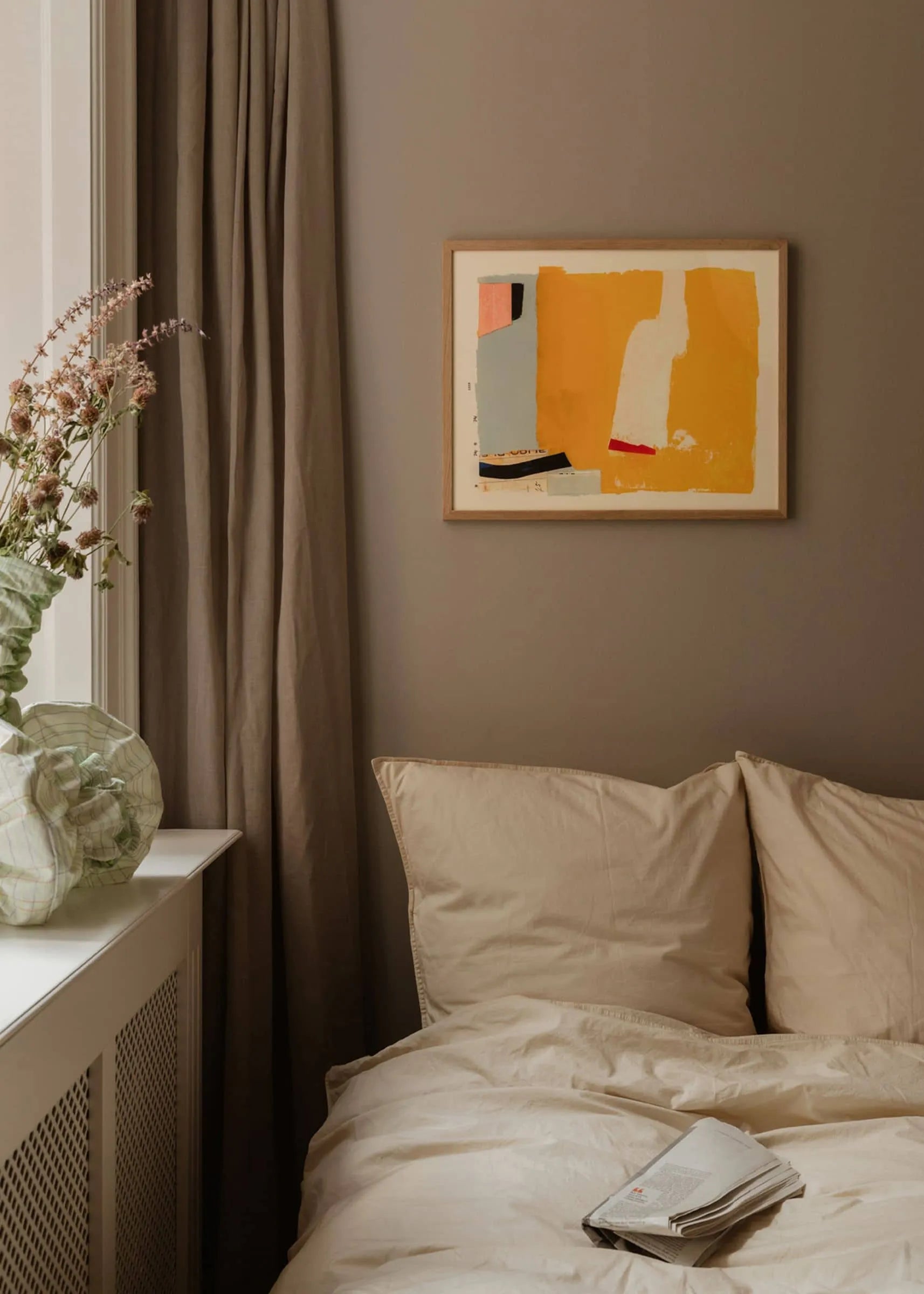A cozy bedroom with beige bedding, taupe curtains, a folded newspaper on the bed, and The Poster Club’s Max Ososki - Jaune (30x40) art print in yellow collage tones on the wall. A glass vase of dried flowers sits on the windowsill.