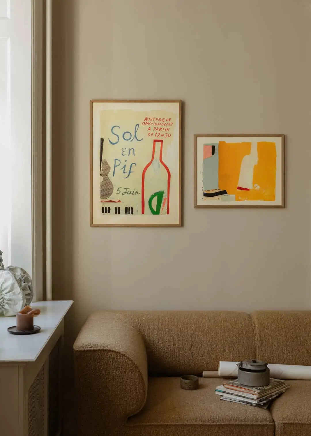 A beige sofa sits against a light tan wall with two framed art prints above it, including Bloom (50x70) by Berit Mogensen Lopez from The Poster Club. To the left, books and decor pieces rest on a white windowsill near a window.