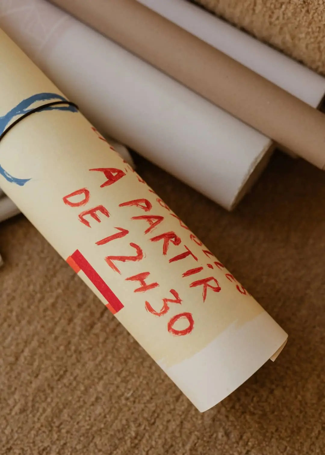 The Poster Club Max Ososki - Sol En Pif (50x70) art print features a rolled paper with handwritten red text A PARTIR DE 12H30 on a beige surface, surrounded by white and brown paper rolls, evoking vintage textile and collage charm.