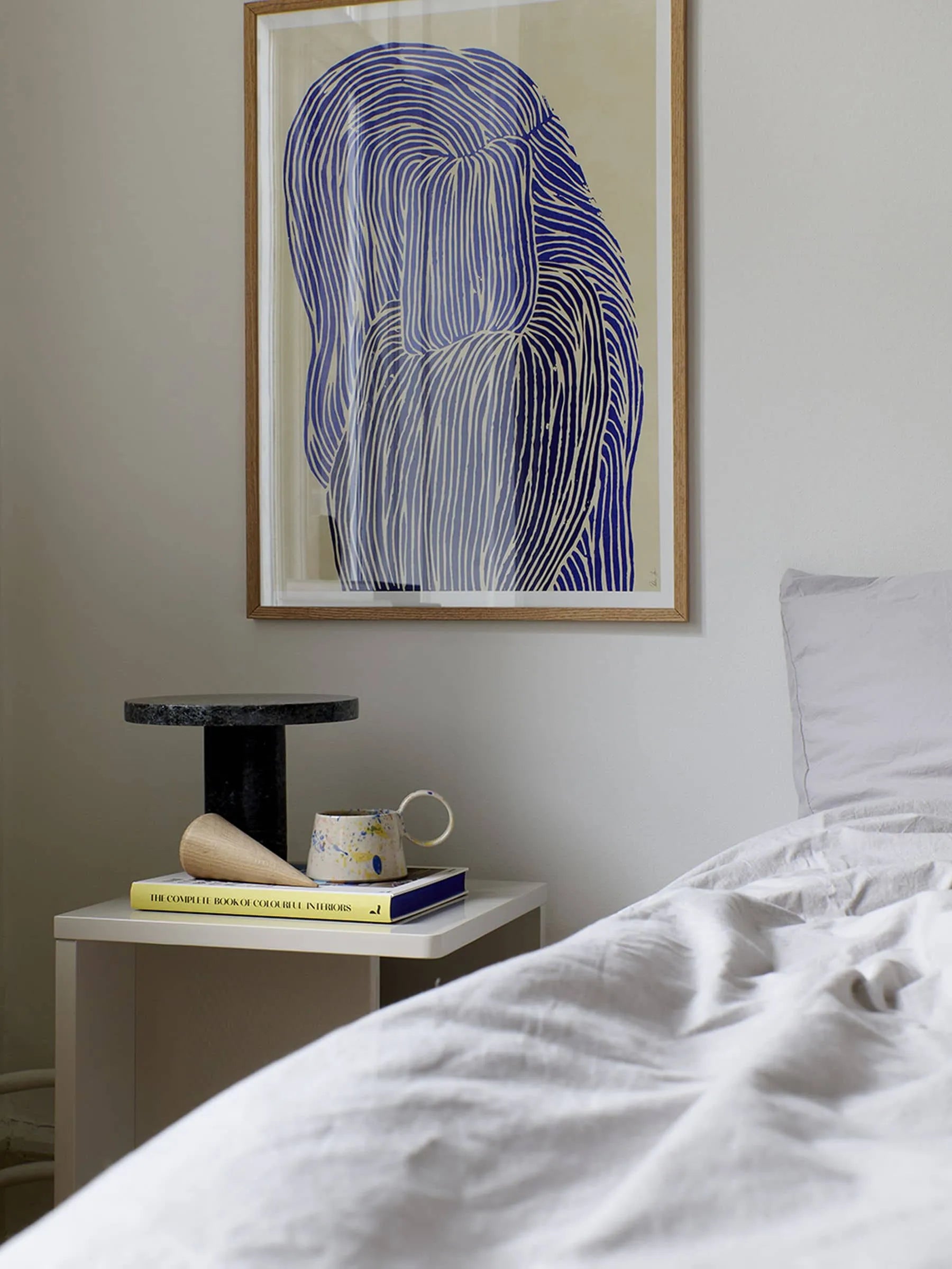 A bedside table with a black pedestal, a mug, and a yellow book sits beside a neatly made bed with white bedding. Above it hangs Rebecca Hein’s Deep Blue (30x40) from The Poster Club, featuring ocean-inspired blue and white art.