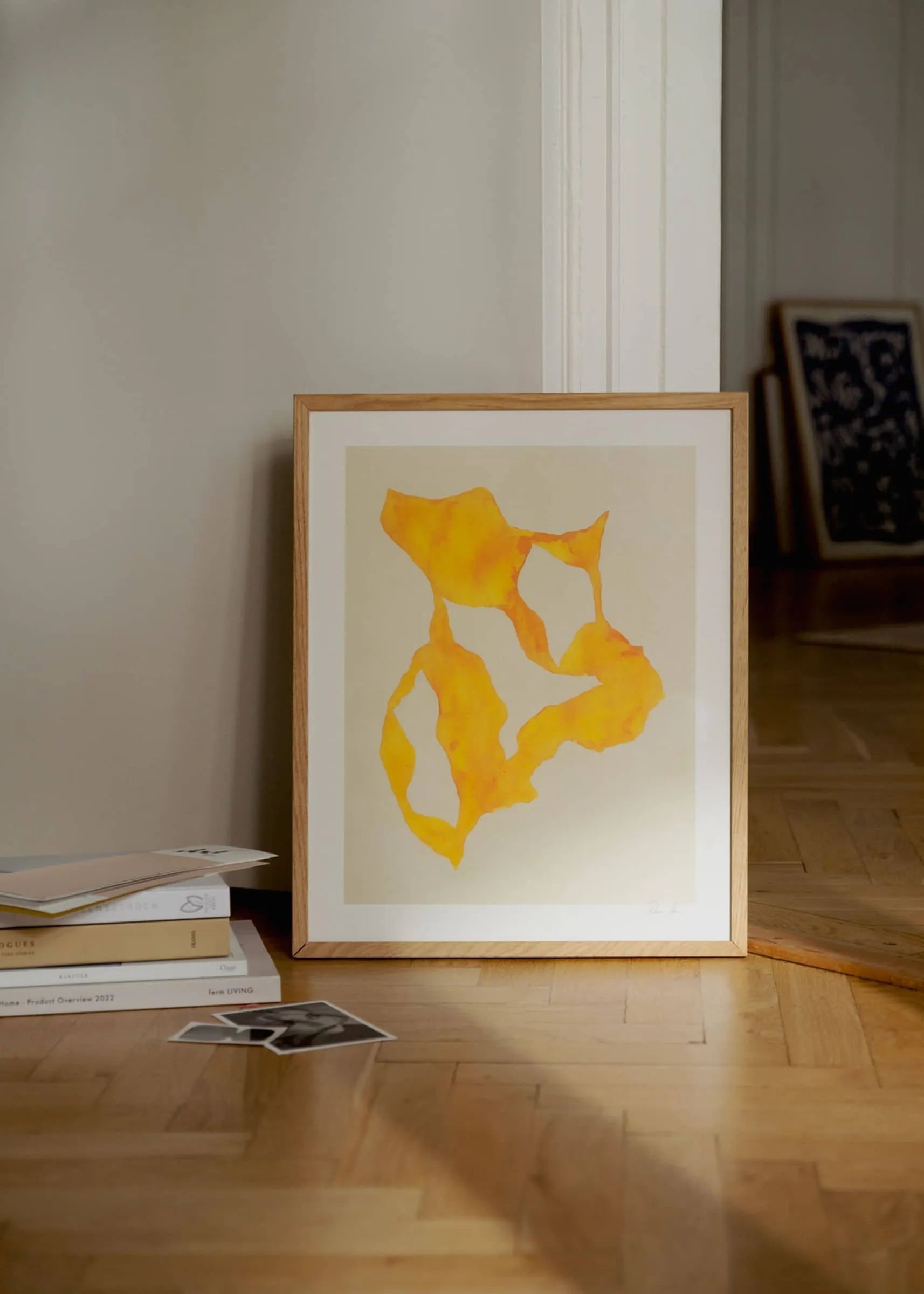 Rebecca Hein - Shadow No. 14 (30x40) by The Poster Club, a framed abstract print with yellow and orange shapes, leans on a wooden floor with stacked books, a black-and-white photo, and another artwork visible in the background.