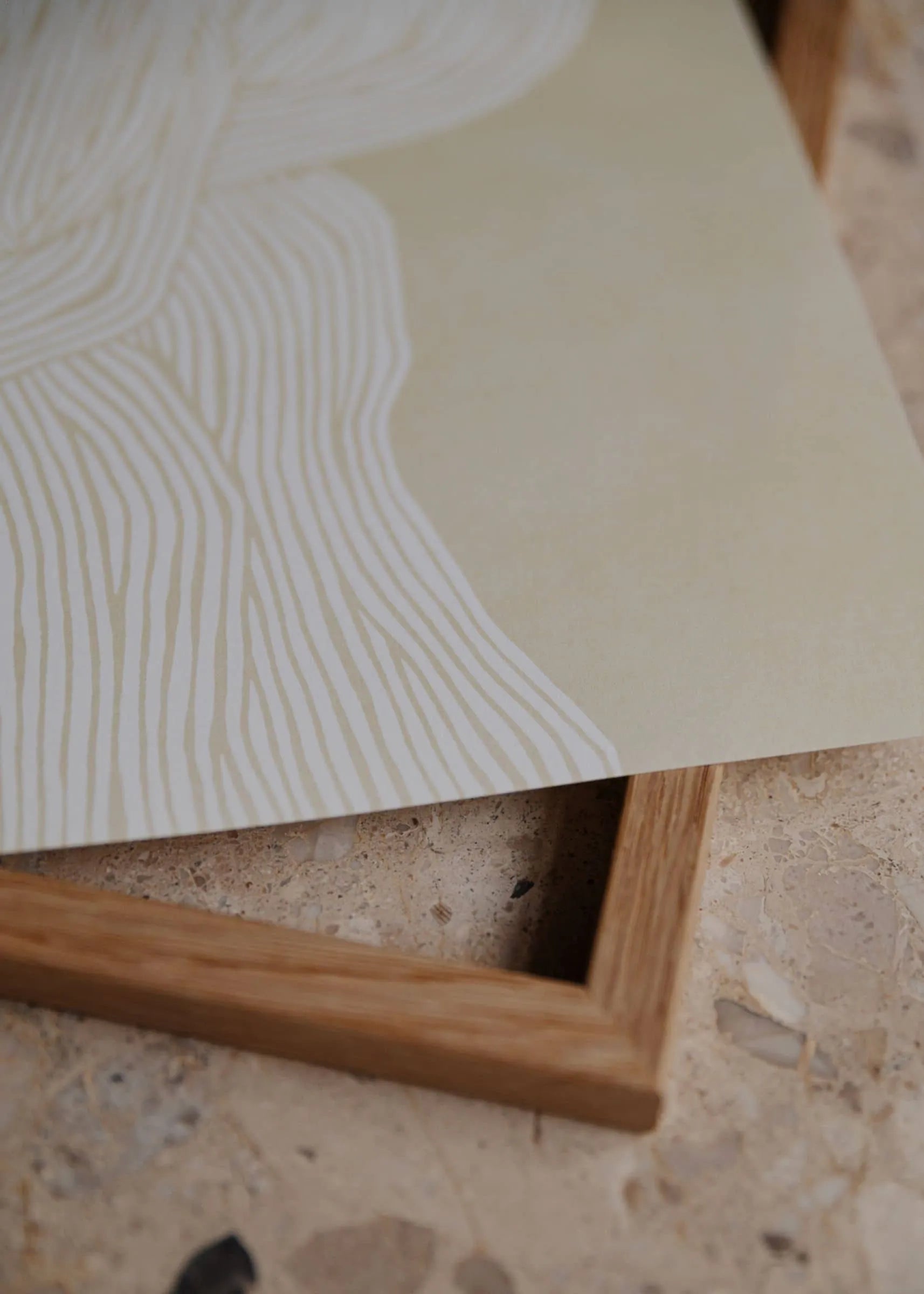 A close-up of The Poster Clubs Rebecca Hein - The Line No. 7 (30x40) art print, featuring white wavy lines on light beige, partially resting on a wooden frame atop a textured stone surface.