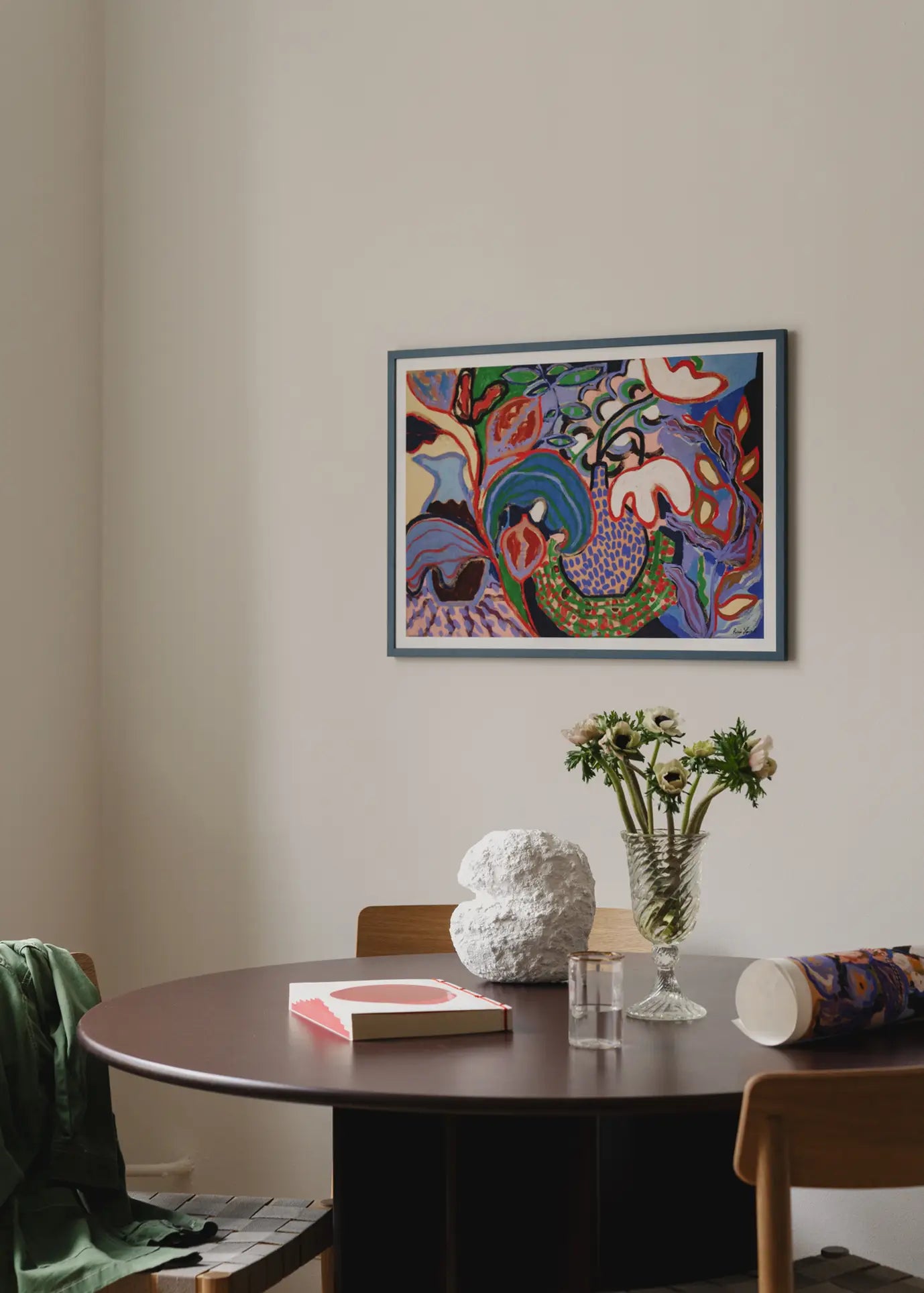 A round dark wood table with a vase of white flowers, a textured white sculpture, a pink book, and The Poster Club Rini Spiel - Felicity (50x70) giclée print sits under a colorful abstract painting. A green jacket is draped over a chair.