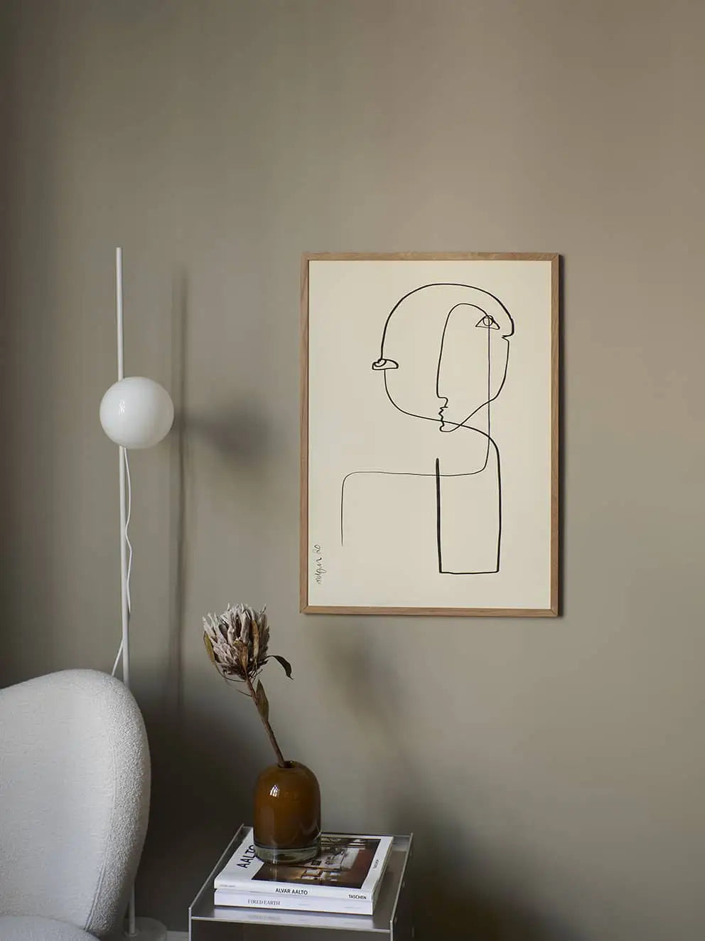 Minimalist room with a neutral wall, The Poster Club Anastasia Krasilnikova - Jaguar 01 (50x70) print in a wooden frame, white floor lamp, white chair, and clear side table with a brown vase of dried flowers and stacked books.