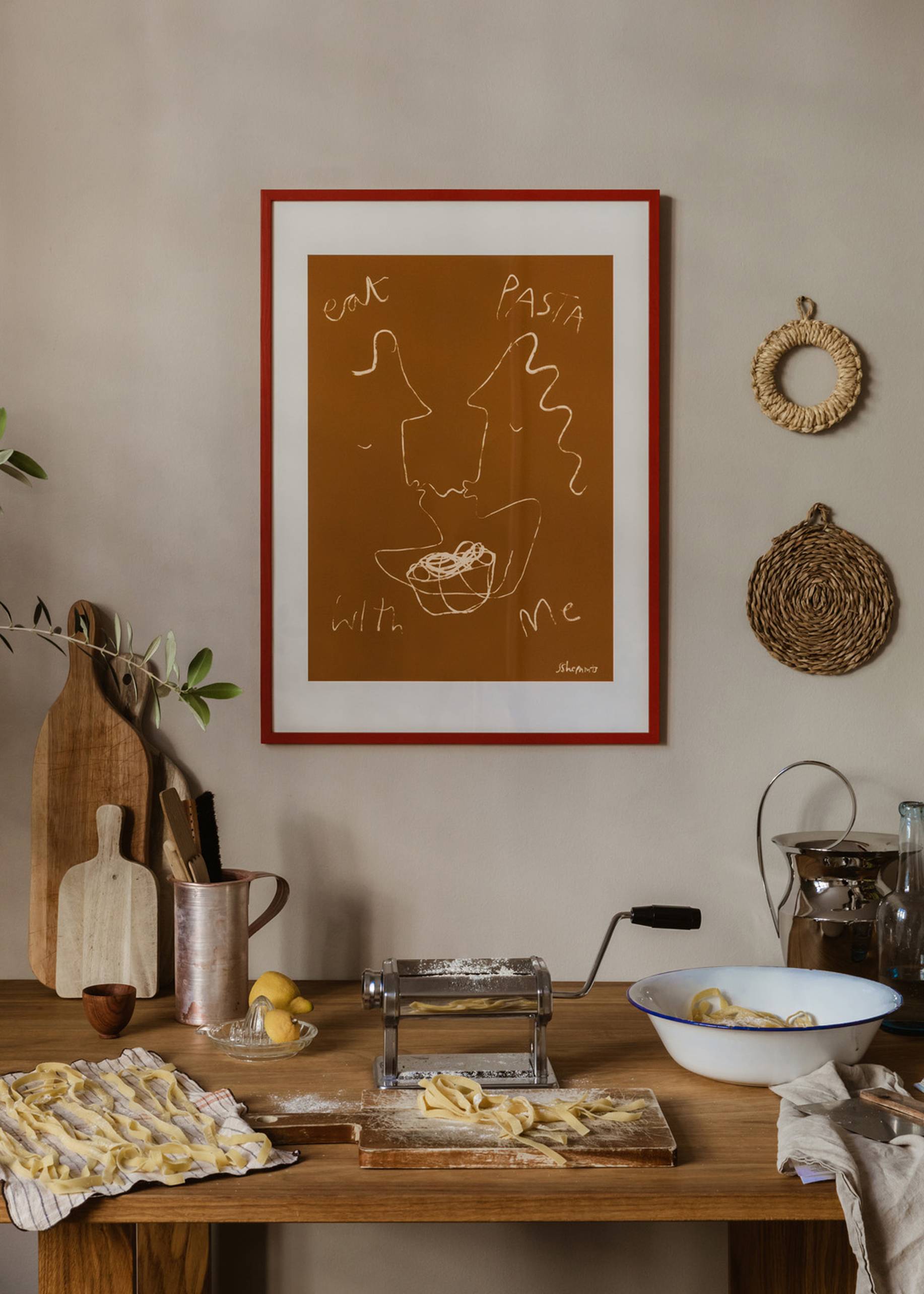 A rustic kitchen features a wooden table with a pasta maker, fresh pasta, eggs, and flour. On the wall hangs The Poster Club Ruby Hughes - Eat Pasta (30x40) art print. Woven decor and cutting boards add charm.