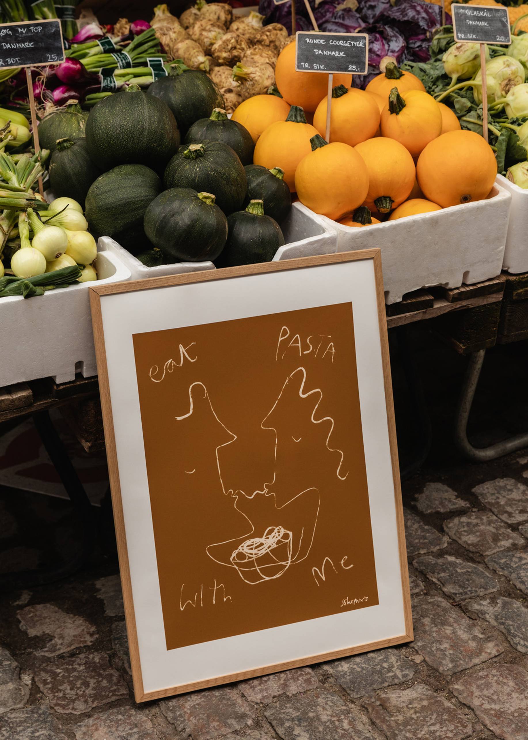 The Poster Club Ruby Hughes - Eat Pasta (30x40) framed art print features a minimalist design with the phrase eat PASTA with me, displayed against market crates of green and yellow vegetables on cobblestone pavement.