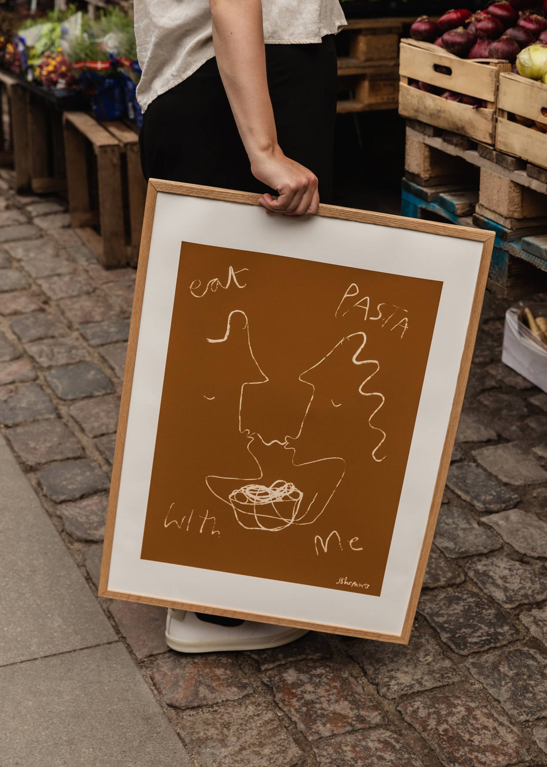 A person holds The Poster Club Ruby Hughes - Eat Pasta (30x40) giclée print, featuring a minimalist line drawing of two faces and pasta with eat pasta with me text on a brown background, showcased at an outdoor market.