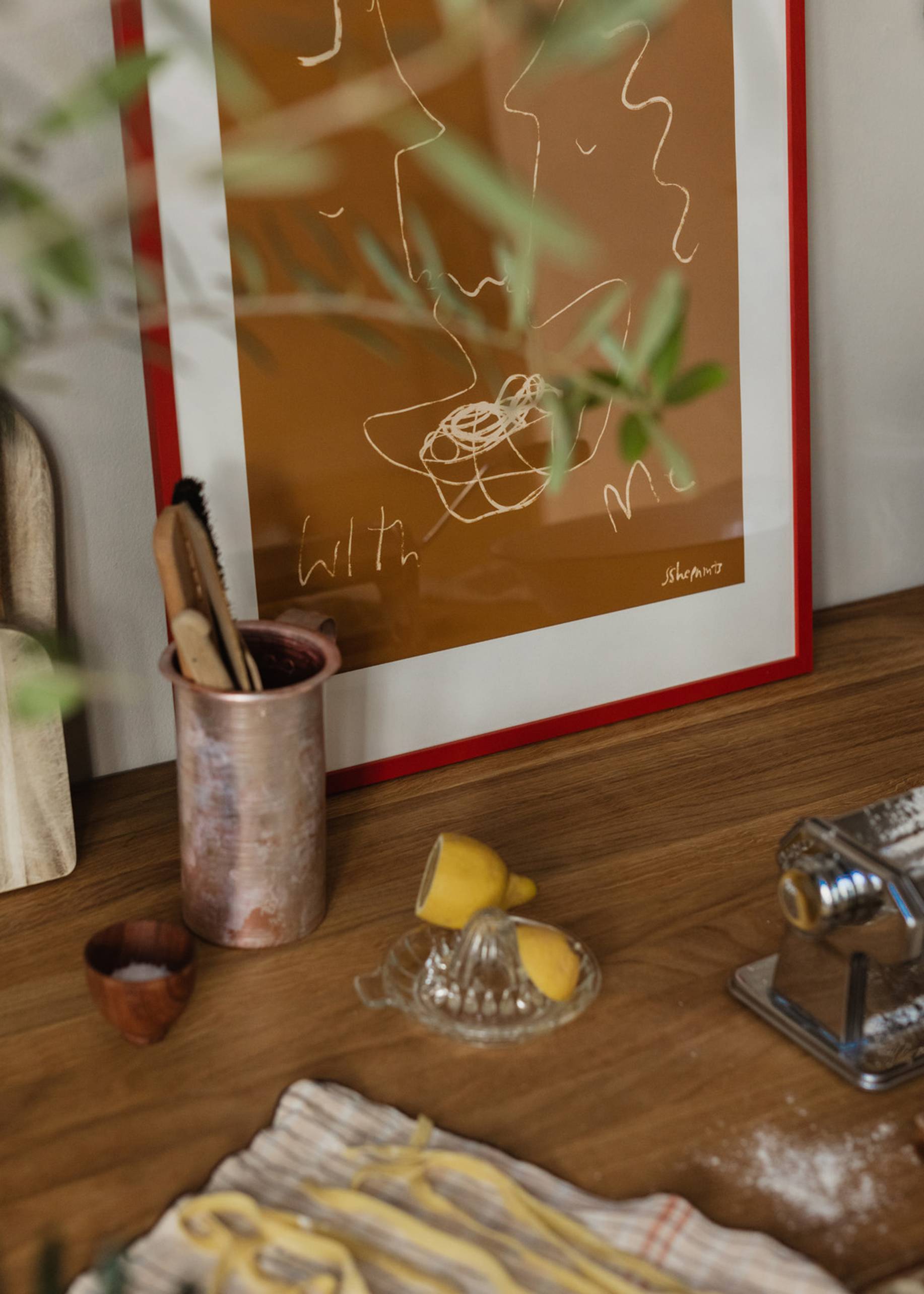 A wooden kitchen counter displays a lemon squeezer, halved lemon, metal pasta maker, kitchen tools in copper, and fresh tagliatelle on a striped cloth. The Poster Club Ruby Hughes - Eat Pasta (30x40) giclée print hangs in the background.