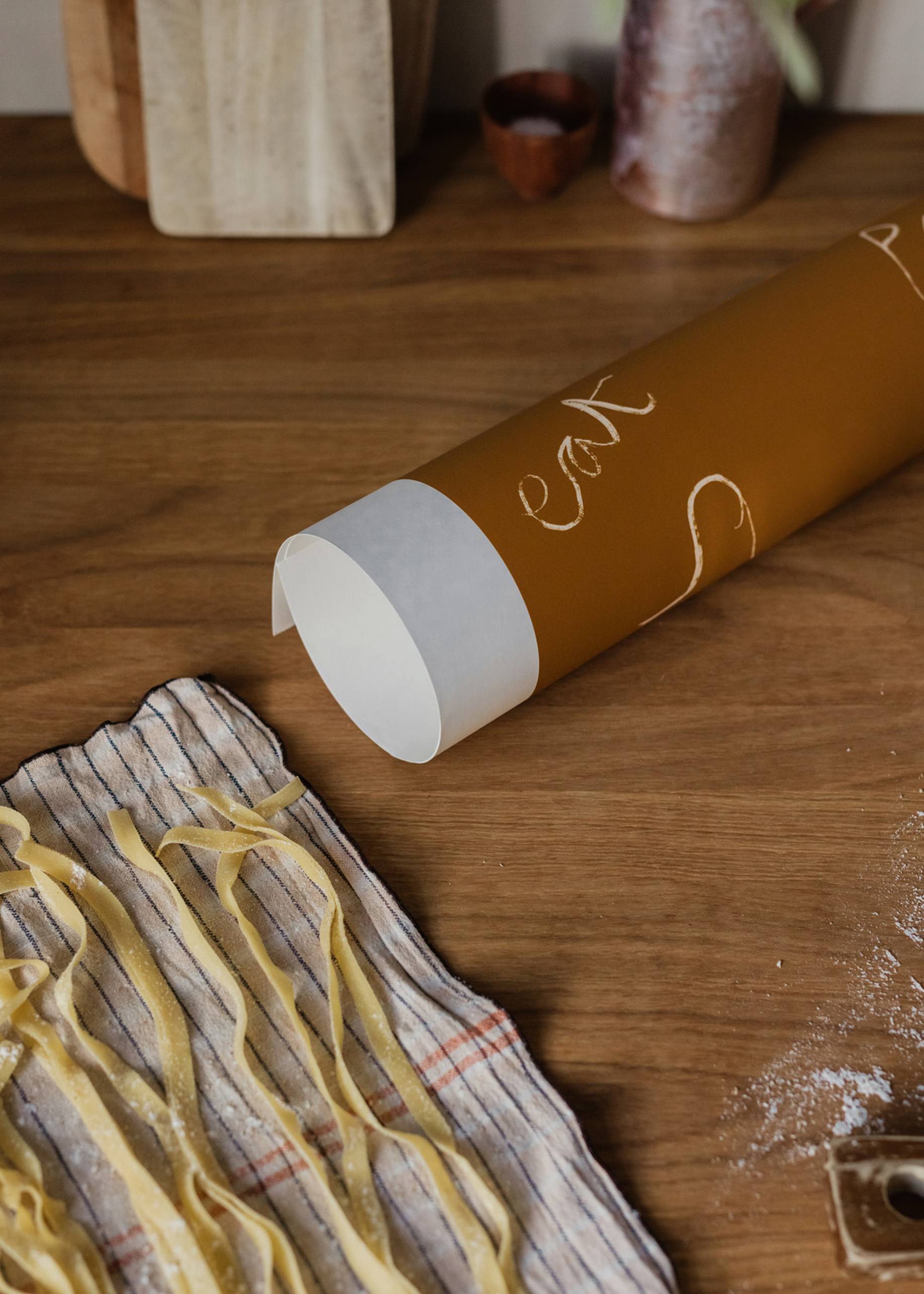 Fresh pasta is arranged on a wooden table, beside handwritten notes on parchment, reminiscent of The Poster Clubs Ruby Hughes - Eat Pasta (30x40) print. Kitchen utensils in the background add to the artistic ambiance.
