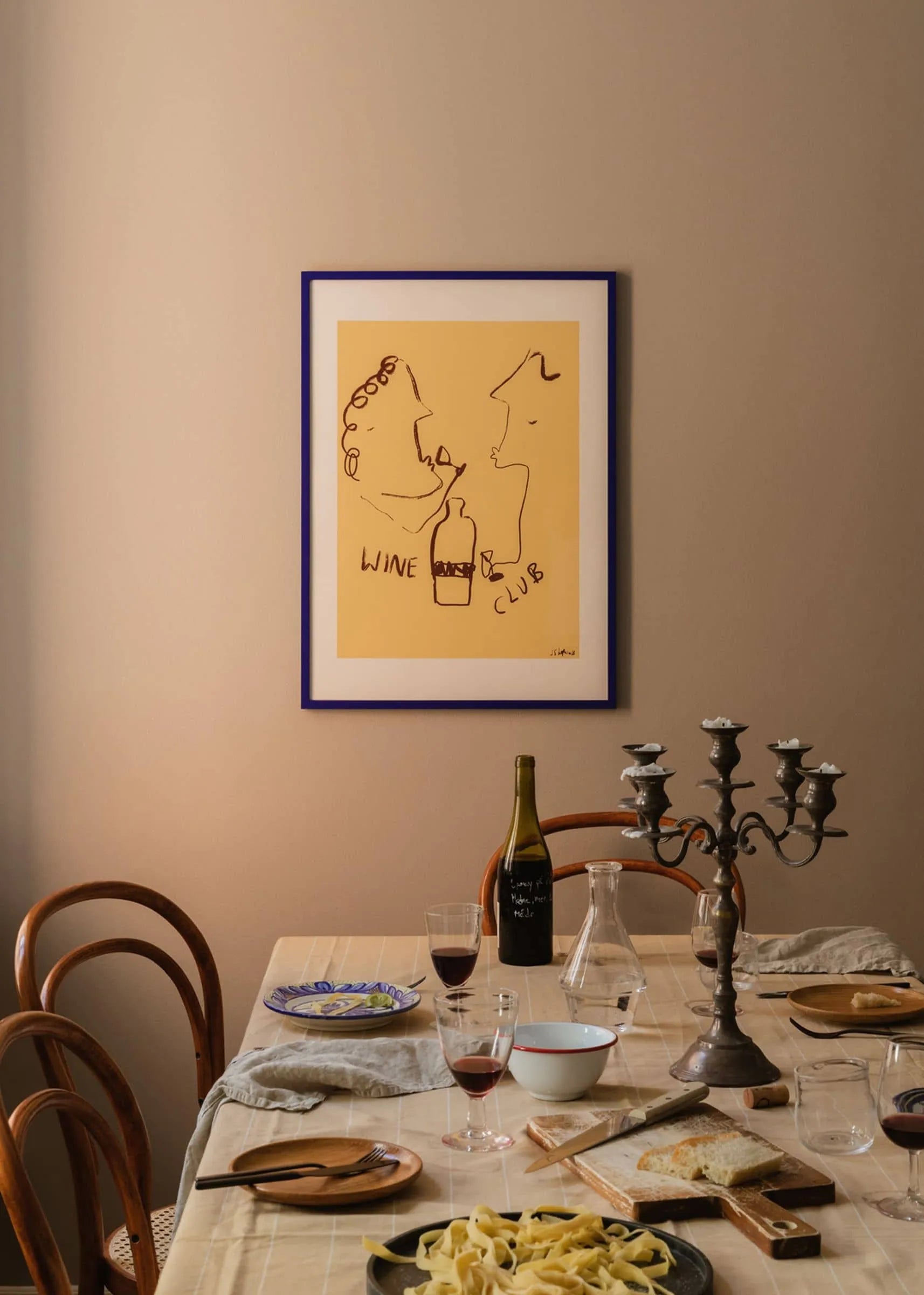 A dining table set with pasta, bread, and wine sits beneath The Poster Club’s “Ruby Hughes - Wine Club (30x40)” giclée print in a blue frame on a beige wall. Wooden chairs and a candelabra add warmth to the inviting scene.