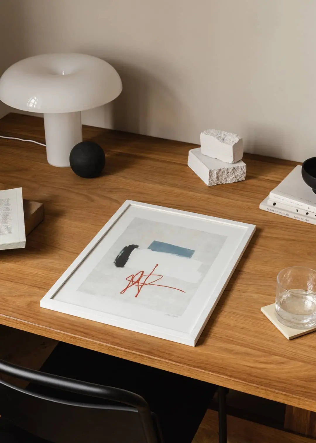 A wooden desk featuring The Poster Club’s “Flowers in Blue Vase” art print by Madelen Möllard (50x70), a glass of water on a coaster, white table lamp, stacked white bricks, an open book, and a black sphere—all neatly arranged.
