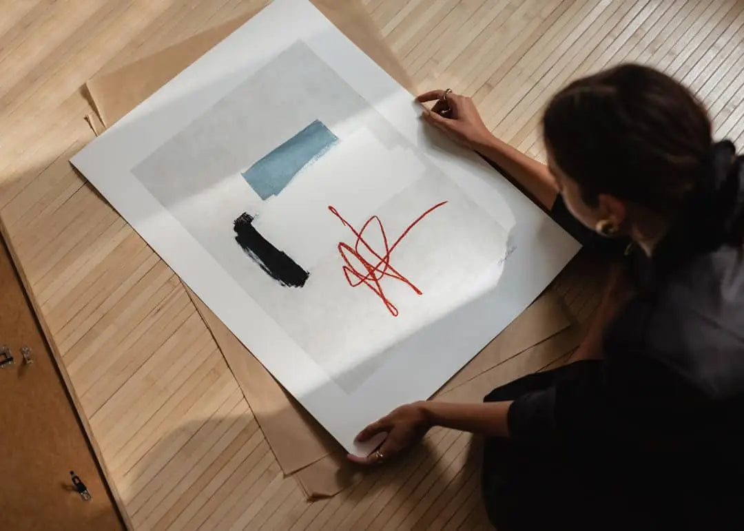 Seated on a wooden floor, a person holds The Poster Club’s Flowers in Blue Vase (50x70) by Madelen Möllard—an abstract art print with blue and black brushstrokes and a red line, surrounded by brown paper sheets.