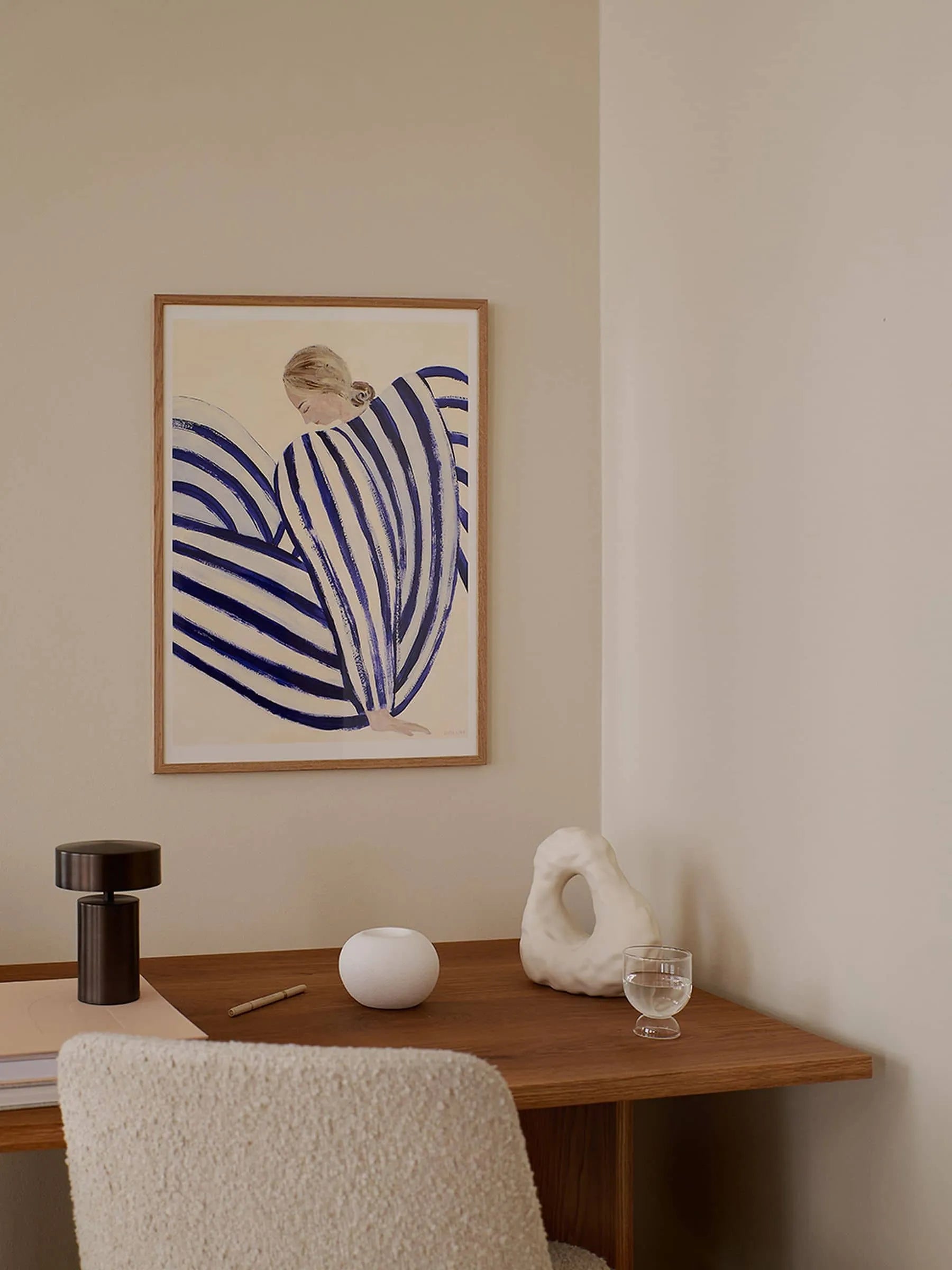 A minimalist workspace features a wooden desk, modern lamp, sculptural object, round container, glass of water, and The Poster Clubs “Sofia Lind - Blue Stripe At Concorde (30x40)” print on the wall.